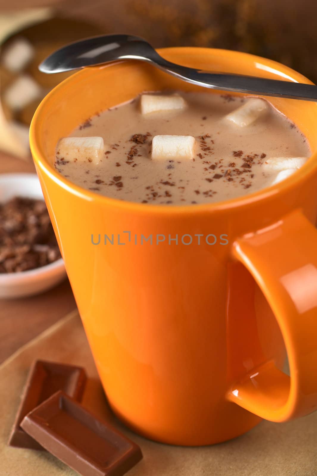 Hot chocolate with marshmallows in orange cup with a teaspoon on the rim and chocolate pieces on the side (Selective Focus, Focus on the marshmallows in the middle of the hot chocolate)