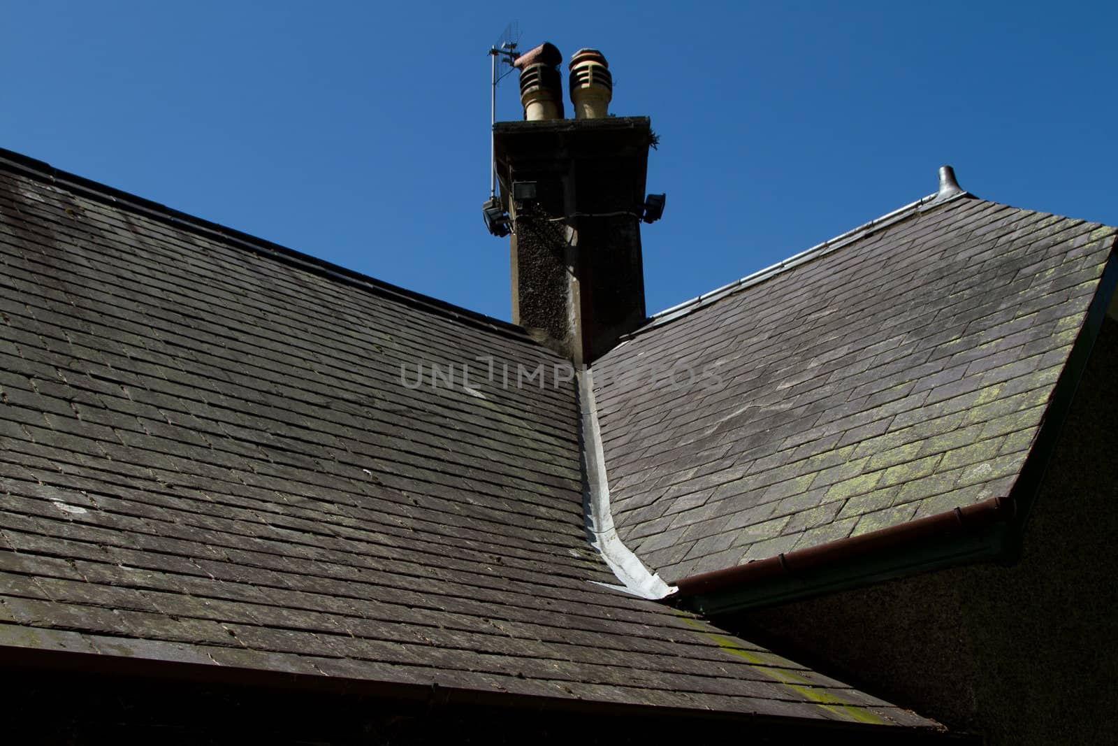 A slate roof with chimney and ridge tiles and a lead valley with green lichen and moss leading off.