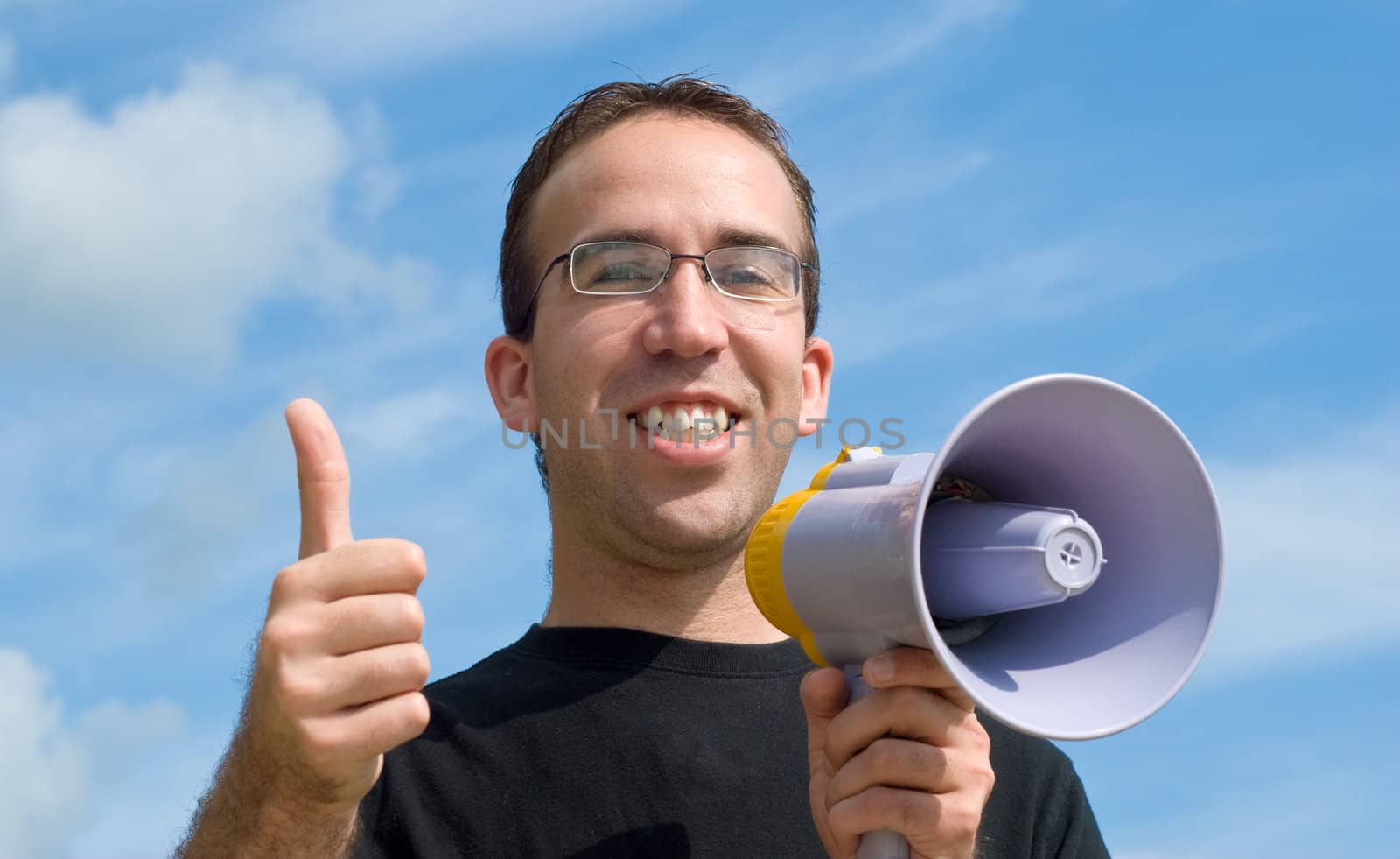 A young man holding a megaphone giving a thumbs up signal, with blue sky and clouds behind him