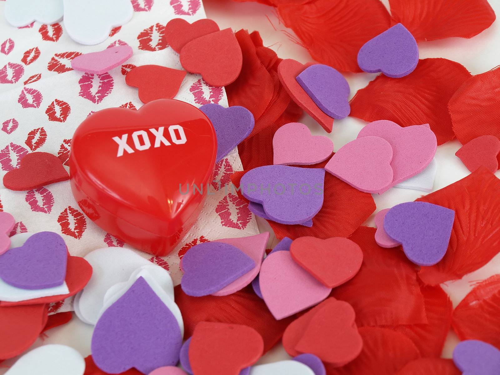 A red plastic heart with XOXO surrounded by foam hearts and lipstick kiss prints. Over white.