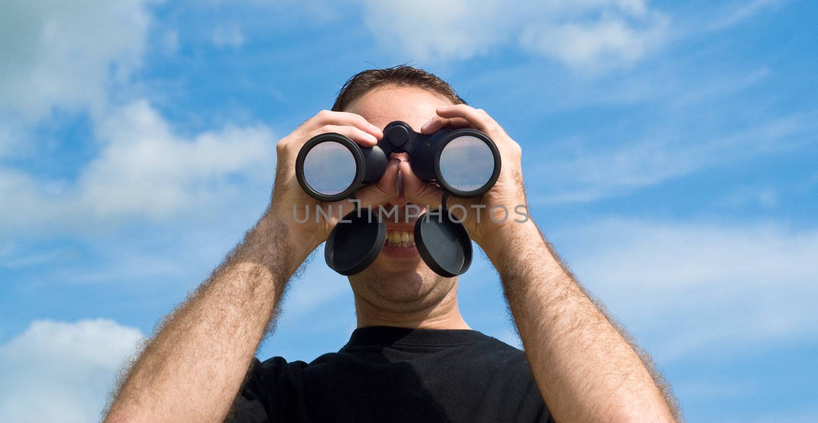 A young man looking at the viewer with a set of binoculars, with a blue sky behind him