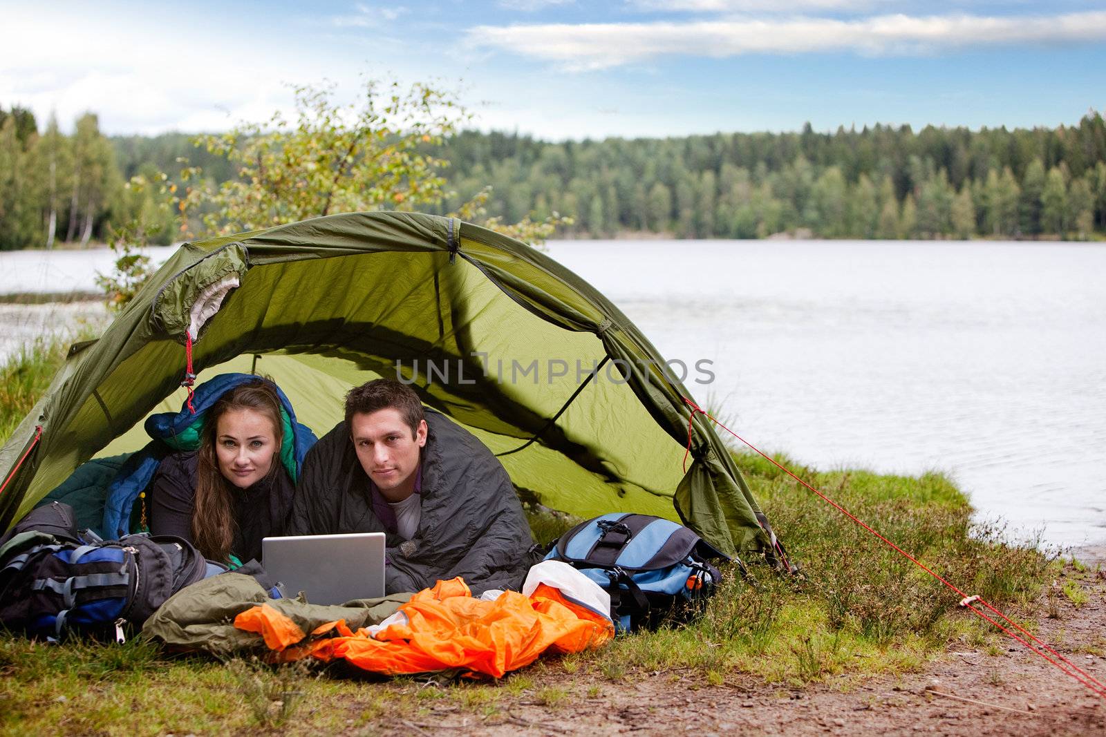 A young couple camping with a laptop near a forest and lake