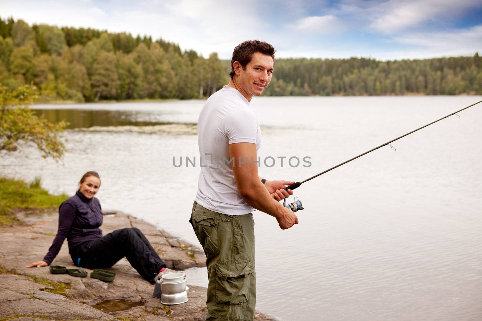 A man fishing on a lake with camping equipment and woman in background