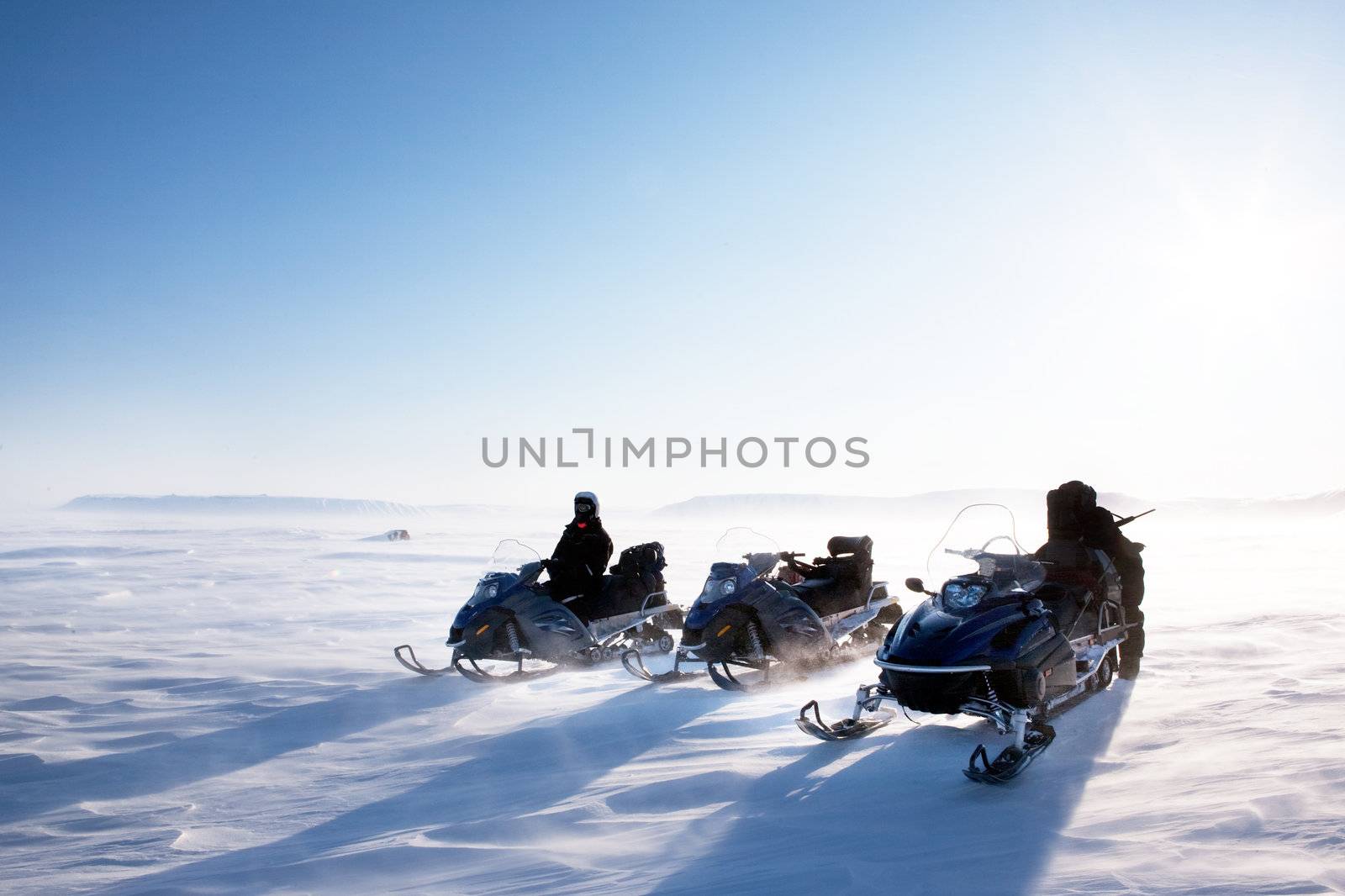 A winter landscape with blowing snow and three snowmobiles.