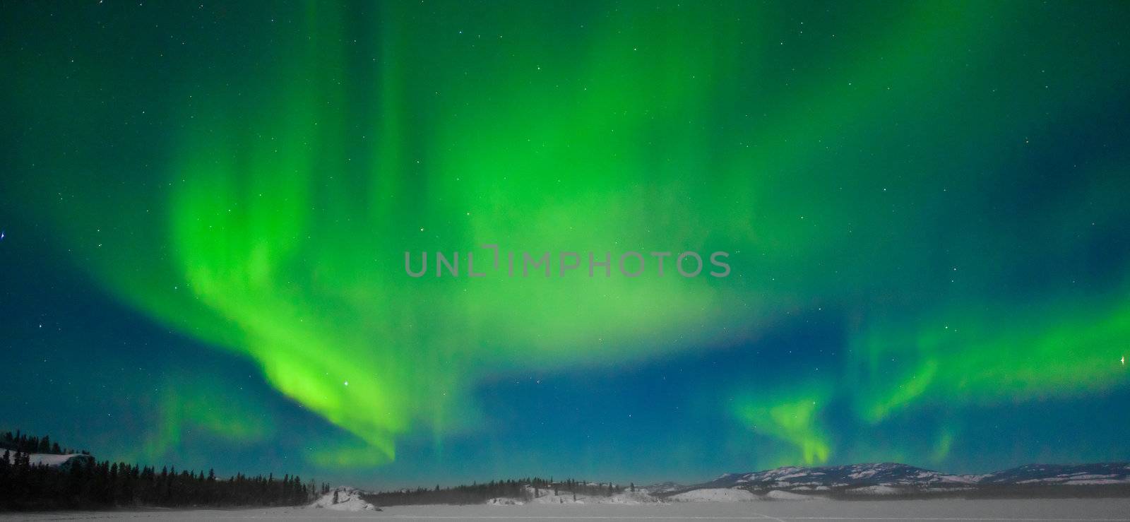 Northern Lights (Aurora borealis) over moon lit snowscape of frozen lake and forested hills.