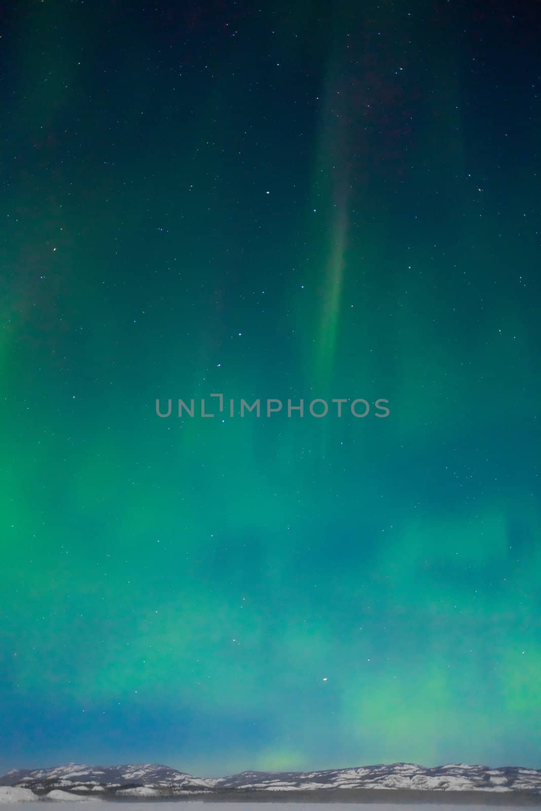 Northern Lights (Aurora borealis) over moon lit snowscape of frozen lake and forested hills.