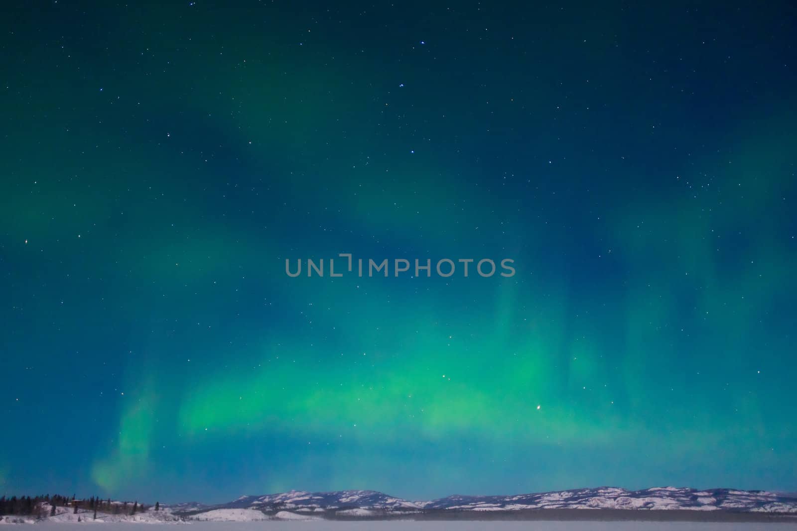 Northern Lights (Aurora borealis) over moon lit snowscape of frozen lake and forested hills.