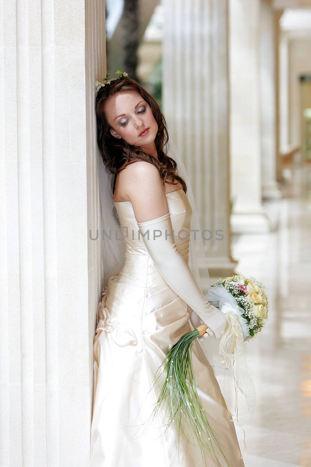 Pretty young adult bride wearing wedding dress and holding bouquet leaning on marble column, thoughtful expression.