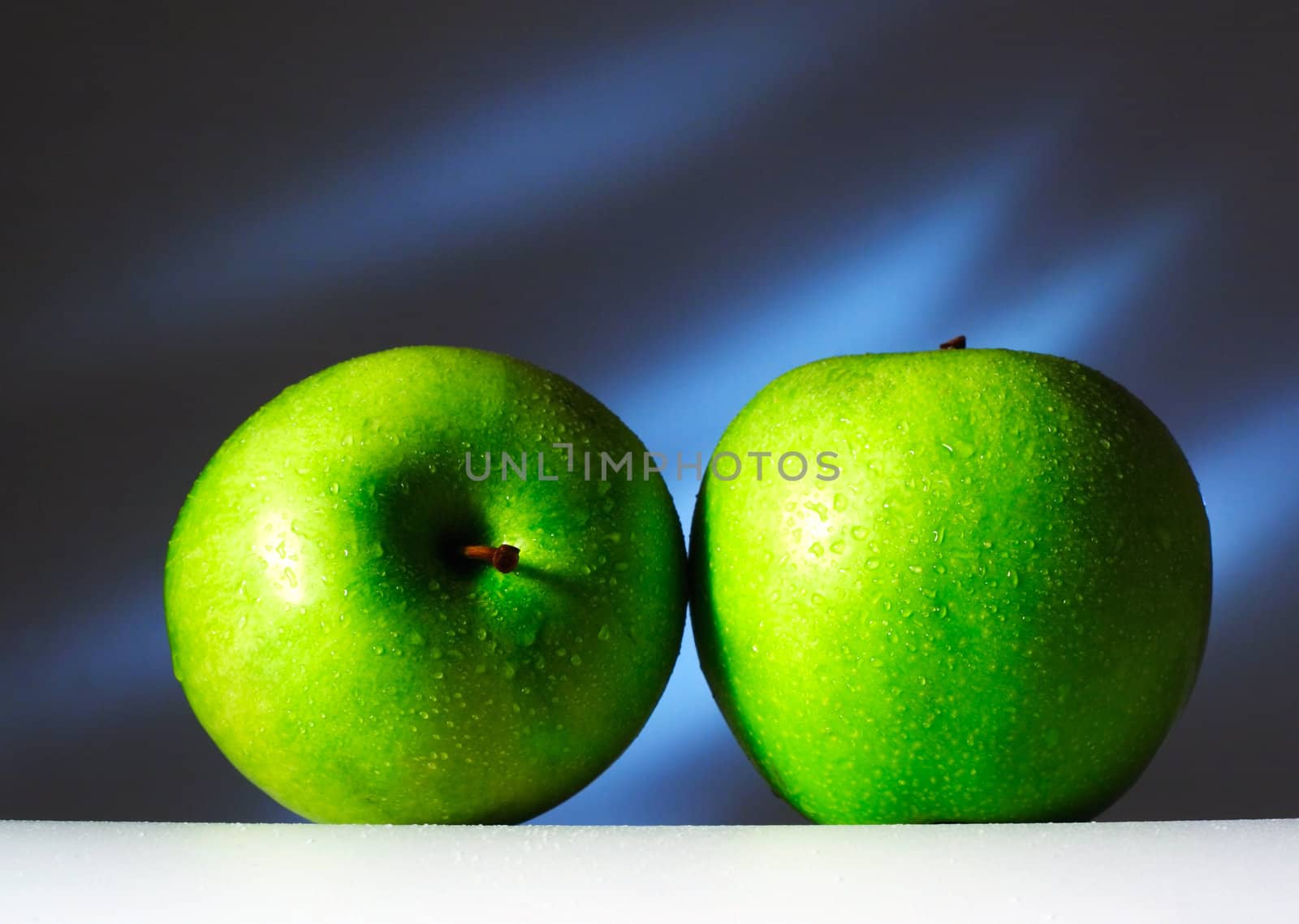 Two green apples on a white surface on a dark blue background. Are covered by water drops