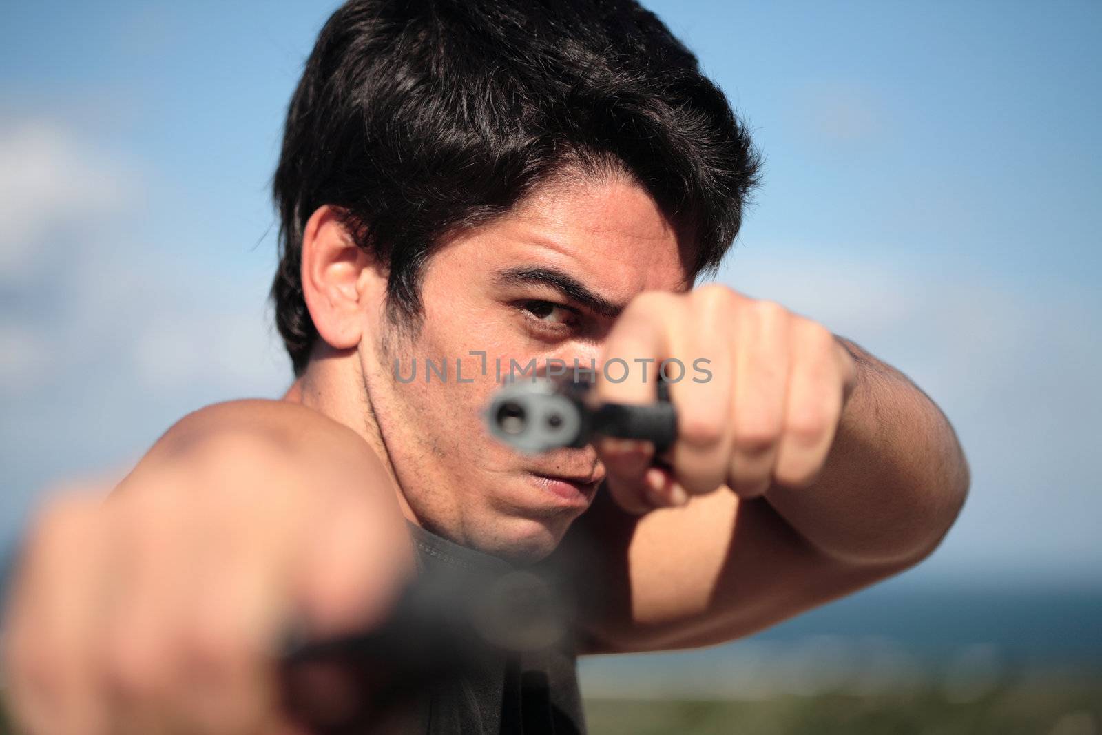 A young, robust man, in his 20's with dark hair pointing 2 pistols to the camera.