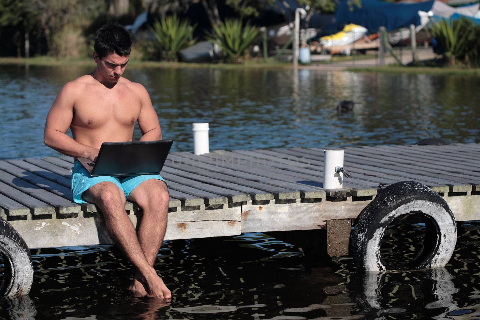 A young man using a laptop on a pier, by the lake, on a warm day, wearing no shirt. 