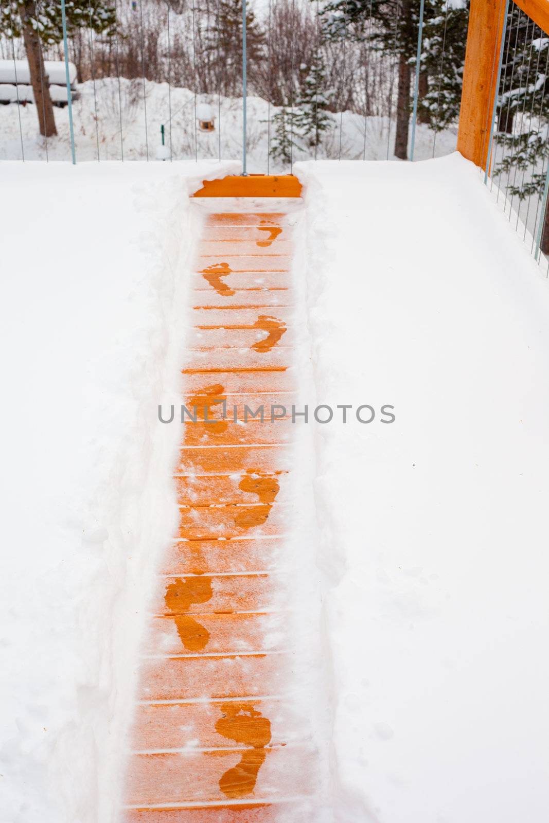 Footsteps of person trying to escape winter by jumping over railing of snow covered wooden deck.