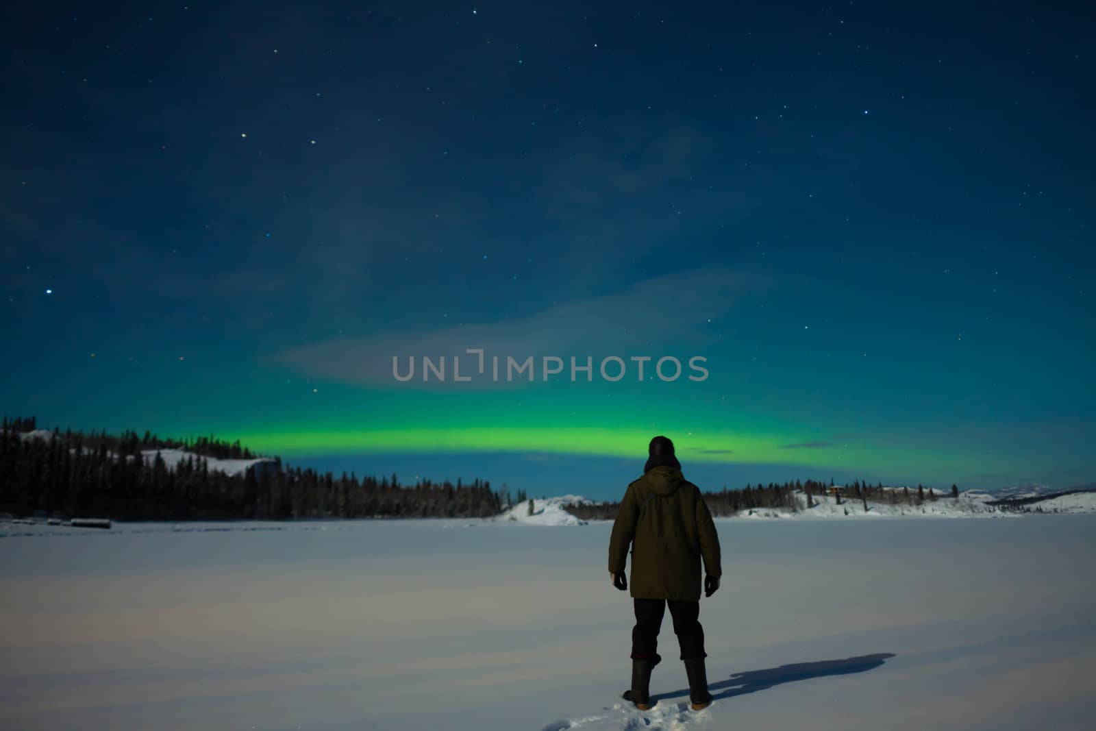 Man watching Northern Lights (Aurora borealis) over moon lit snowscape of frozen lake and forested hills.