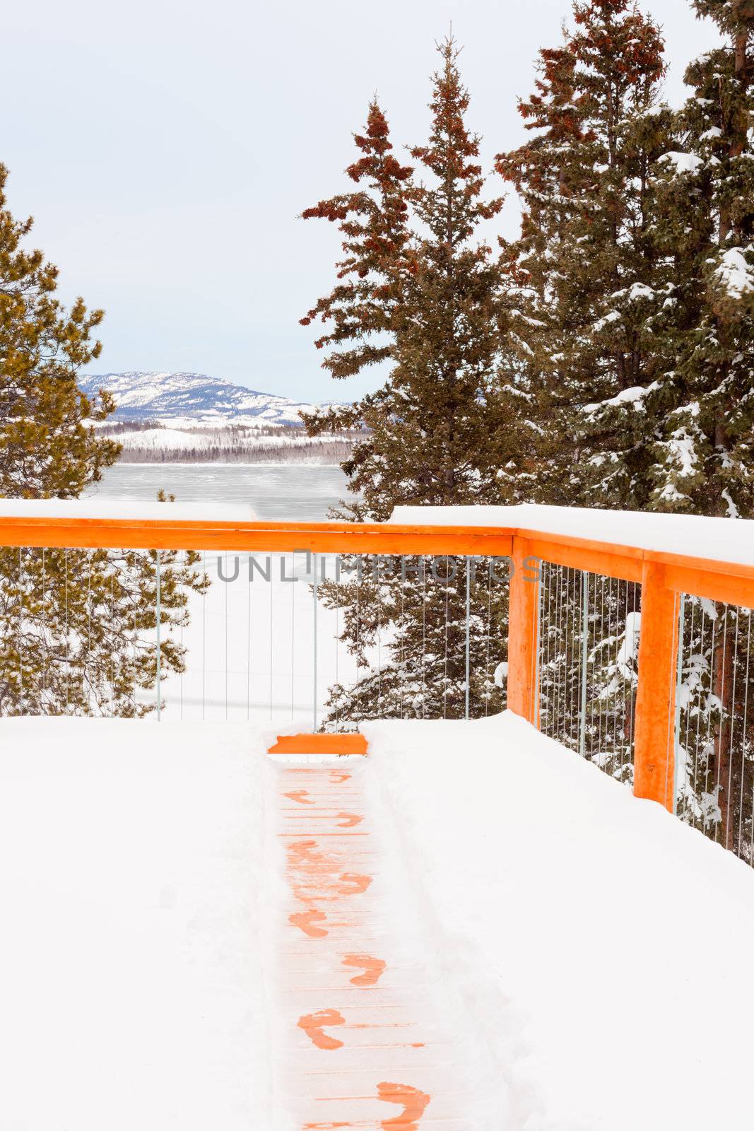 Footsteps of person trying to escape winter by jumping over railing of snow covered wooden deck.