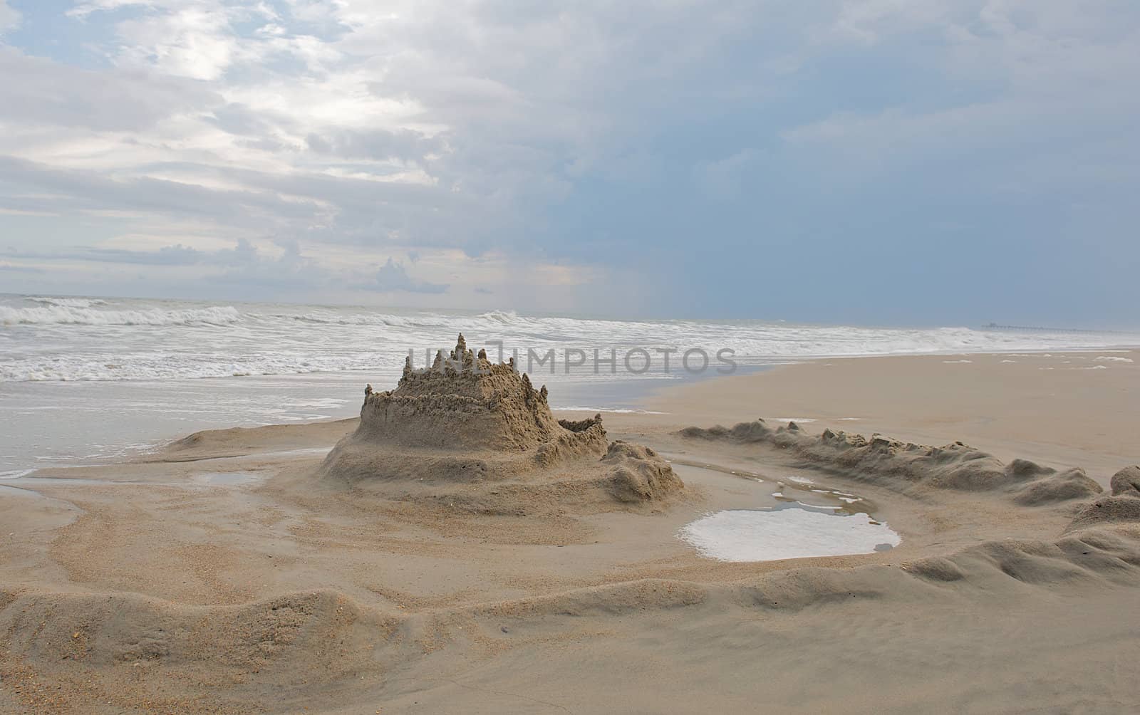 Landscape image of a sand castle with a moat around it.  Ocean and summer sky both included in image.