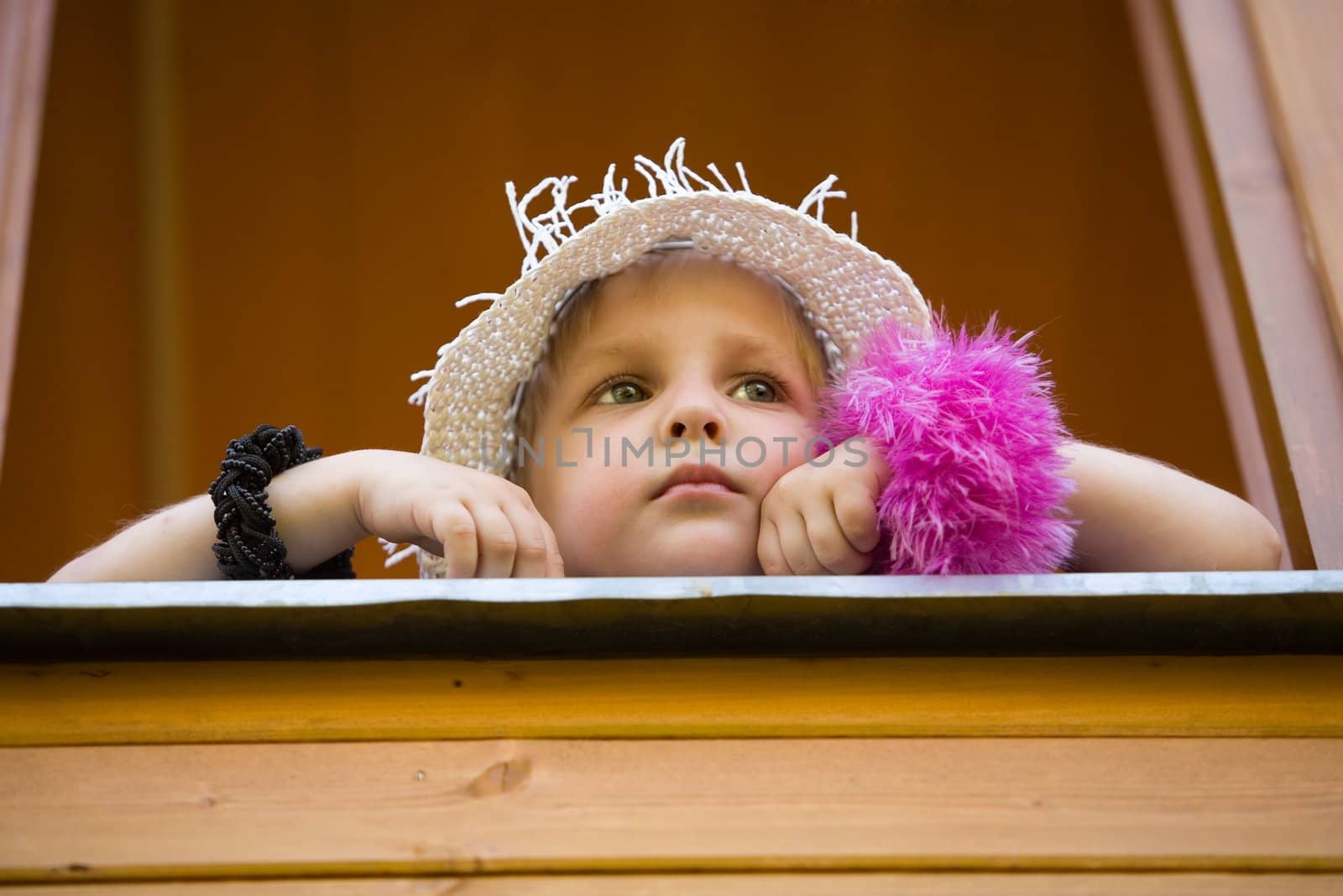 The little girl in a summer hat looks out of a window of a wooden country house
