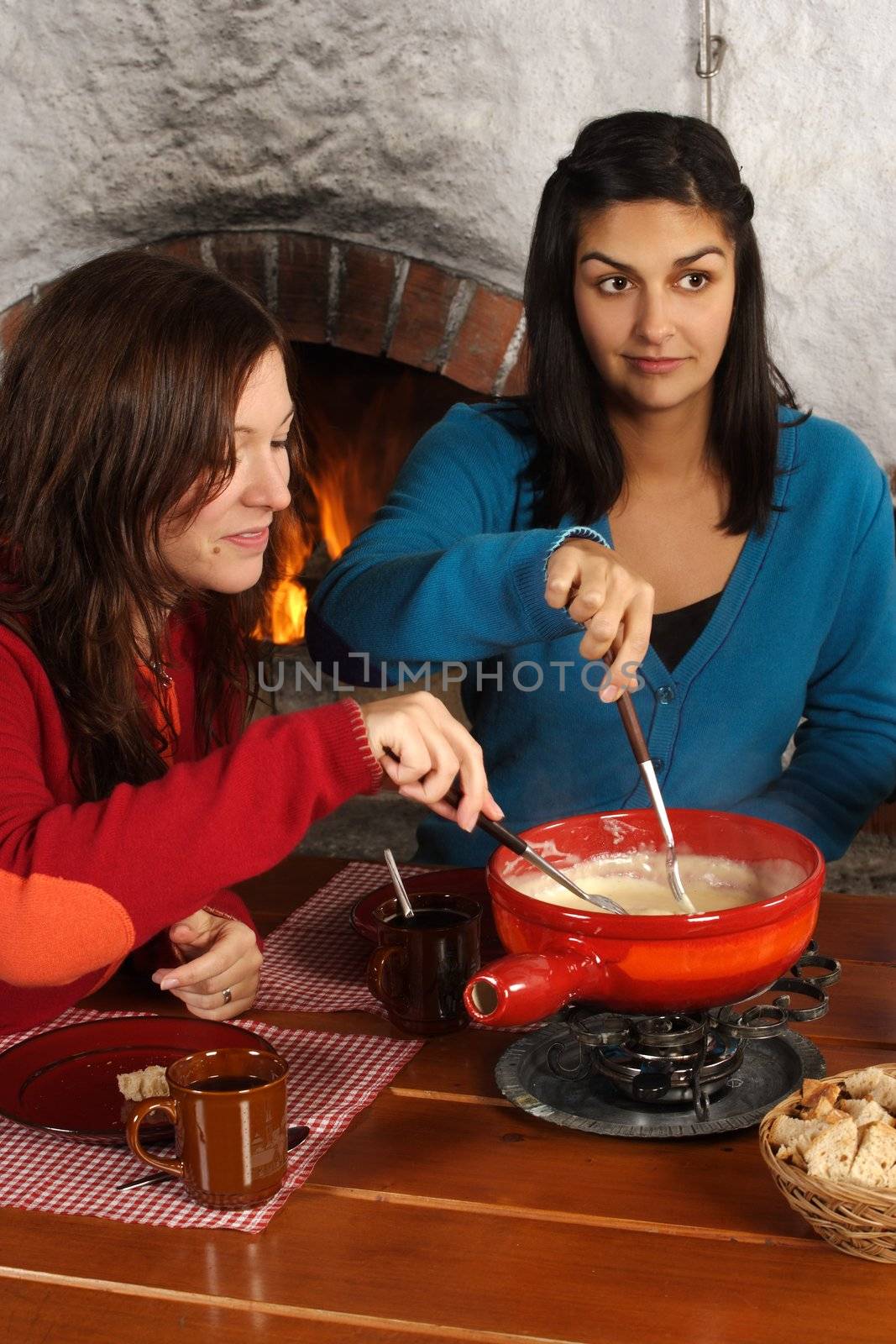 Photo of two beautiful females dipping bread into the melted cheese in a fondue pot. Focus is on girl on the left.