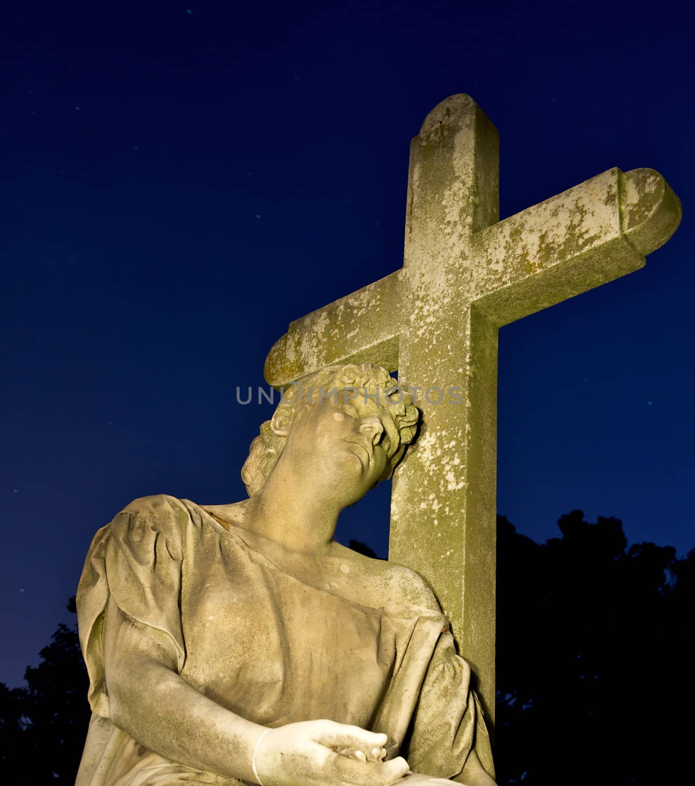 Cemetary headstone of a man resting his head against a cross surrounded by a dark blue night sky.