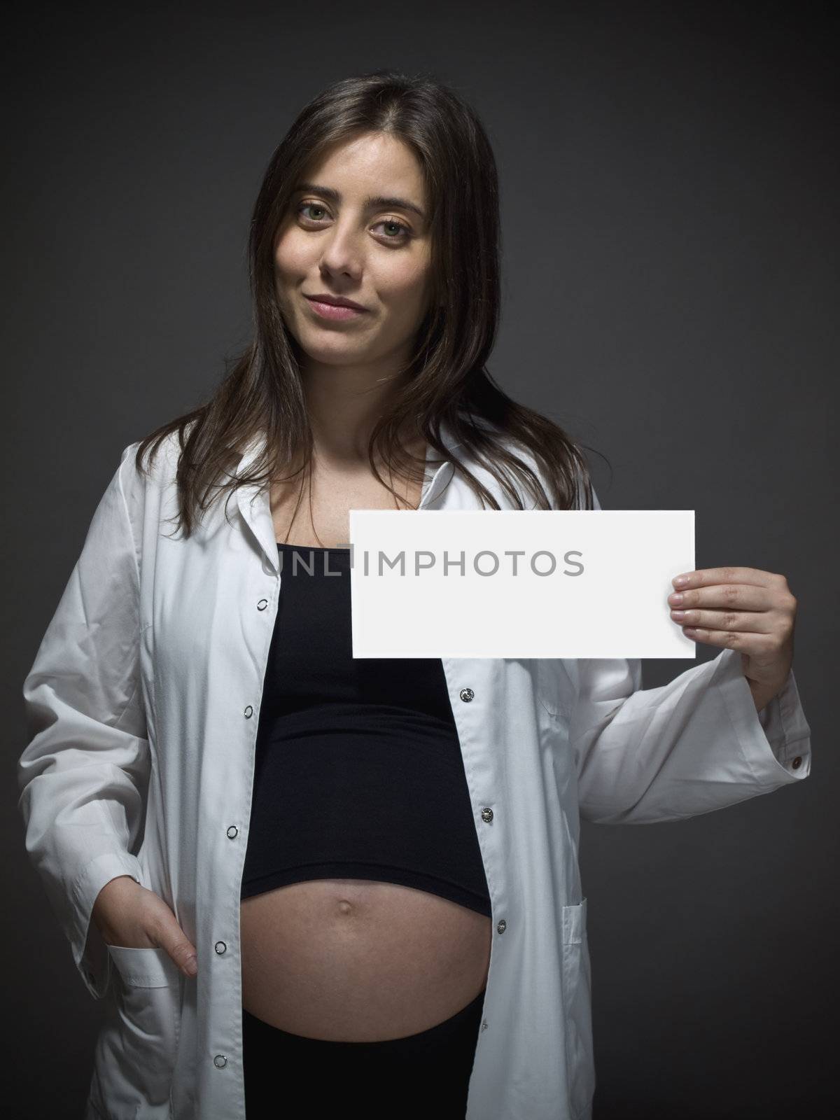 Pregnant female doctor holding a white card over gray background.