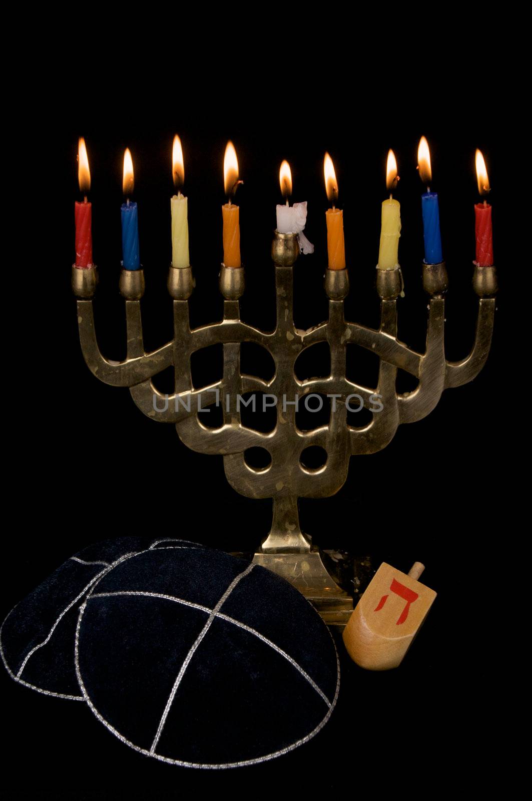 A menorah with 9 burning candles is seen with two yarmalkas and a dreidel, isolated on black