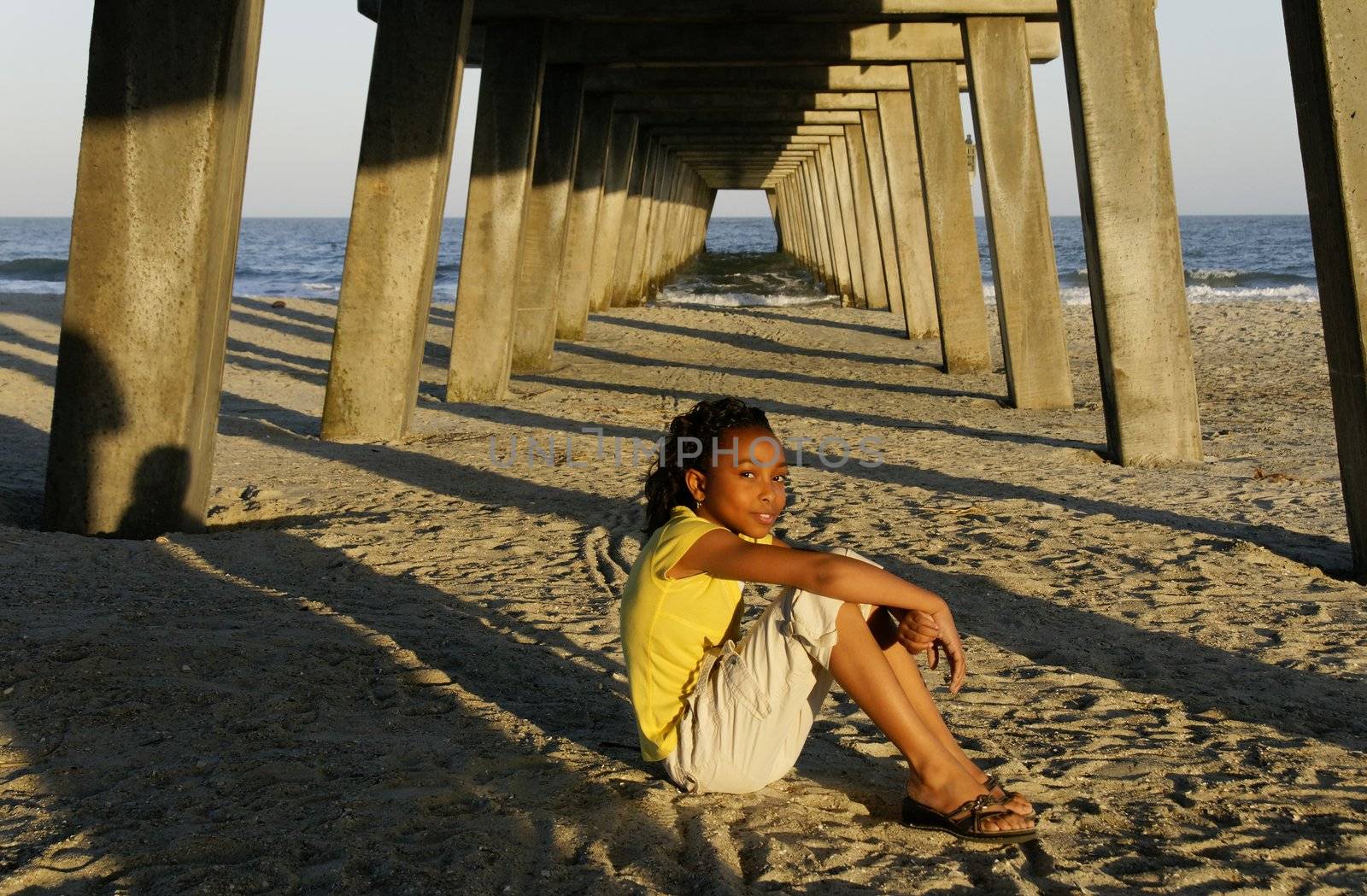 An african-american girl sitting on the sand at the beach with a pier in the background.