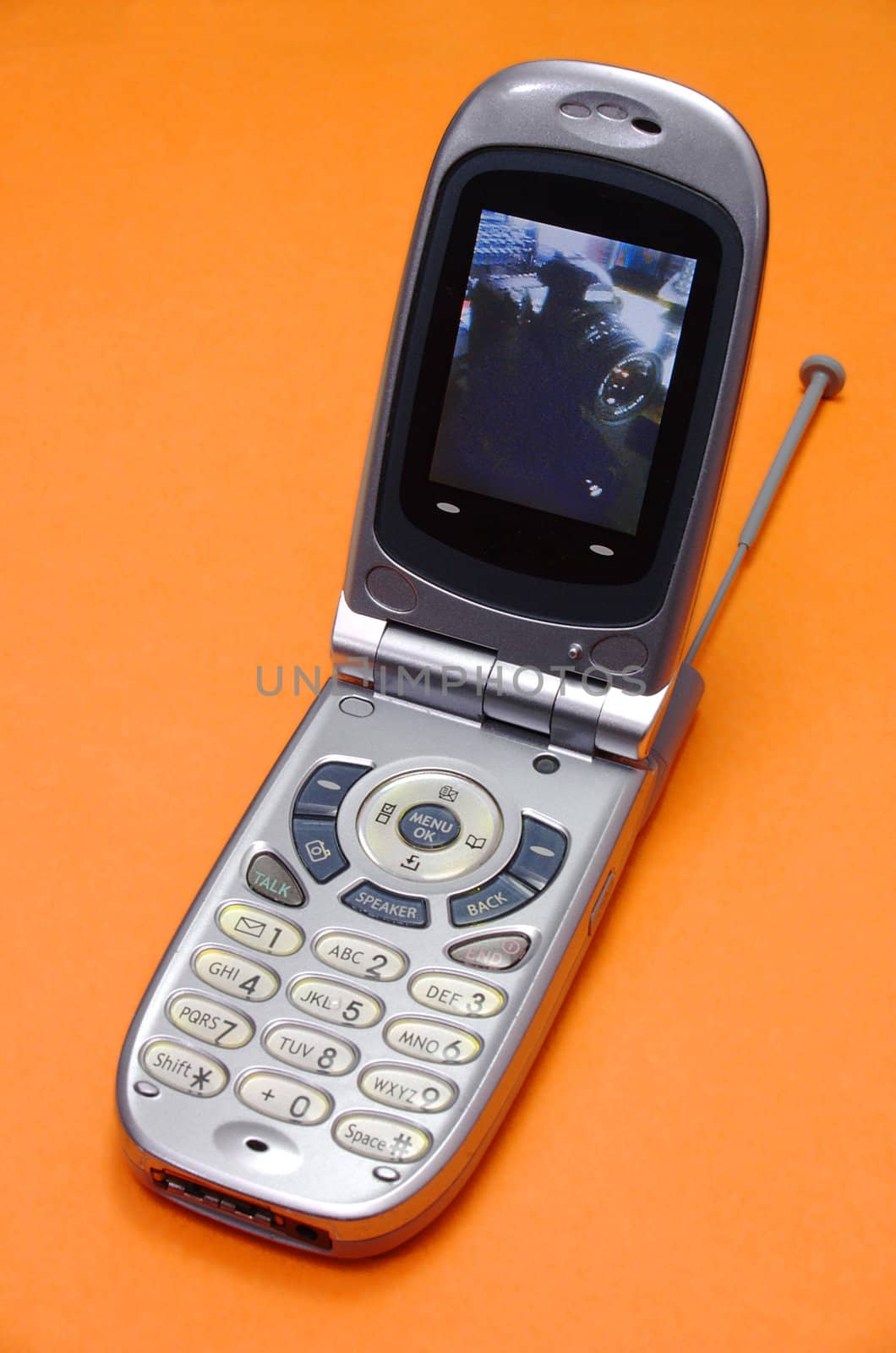 A silver, flip-type cellphone, opened and showing a photo of an SLR camera on it's screen. The phone is laid against a bright orange backdrop.
