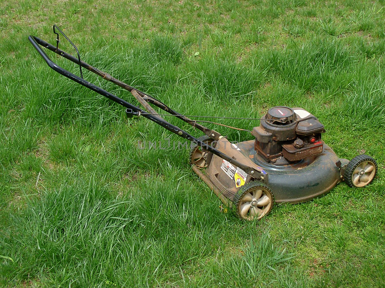A dusty gasoline-powered push lawnmower stands in long grass which is badly in need of mowing.