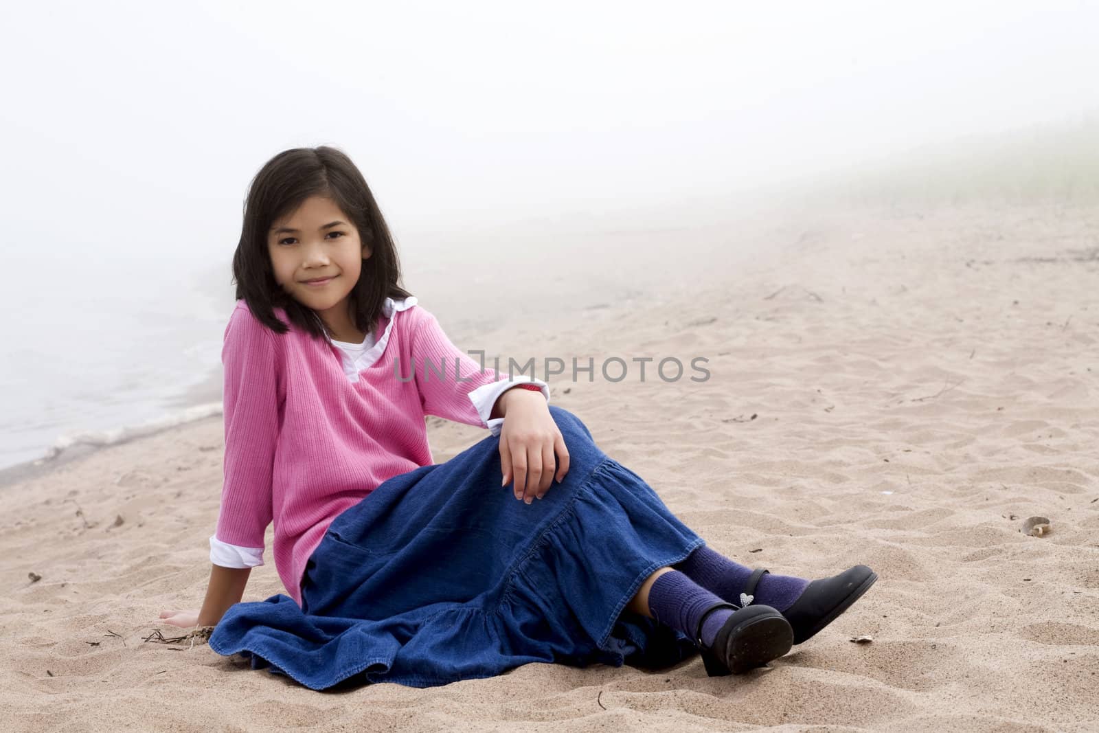 Young girl sitting on beach by lake on misty foggy day