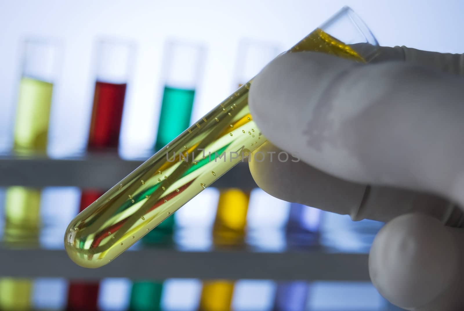 Lab personnel handles test tube, studio shot; line of test tubes containing different colored liquids at background

