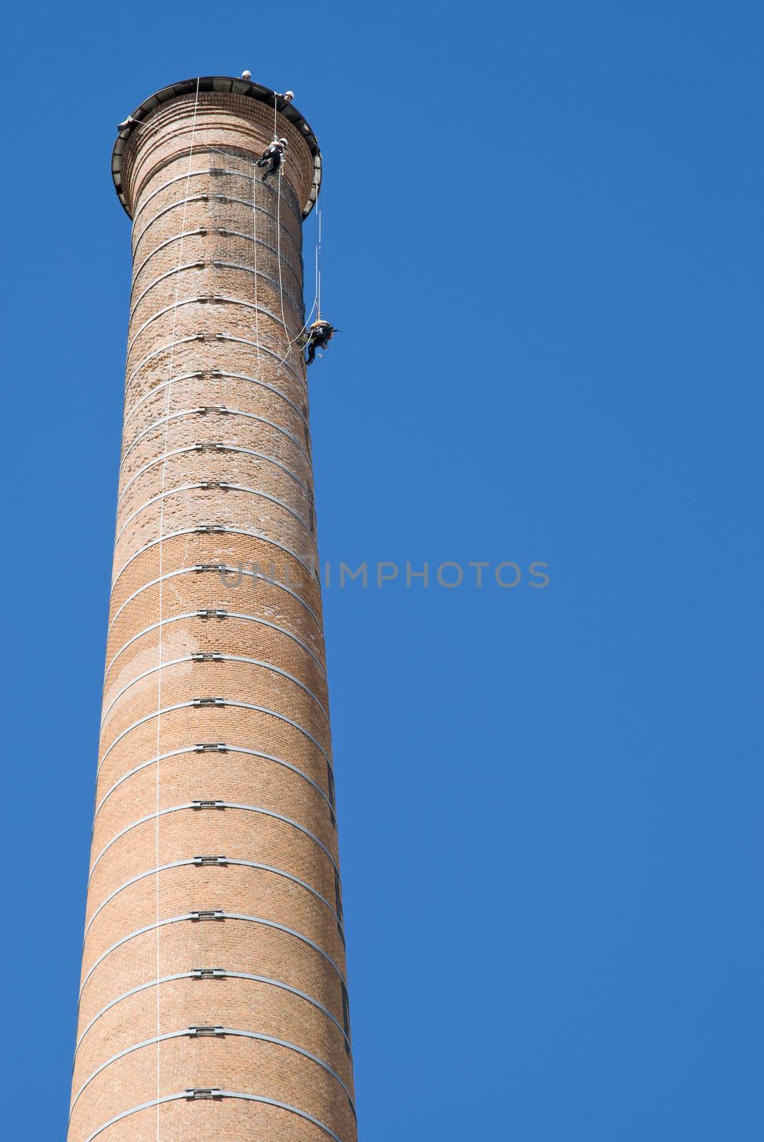 Huge Industrial Chimney with unrecognizable workers hanging from it. Blue sky.
