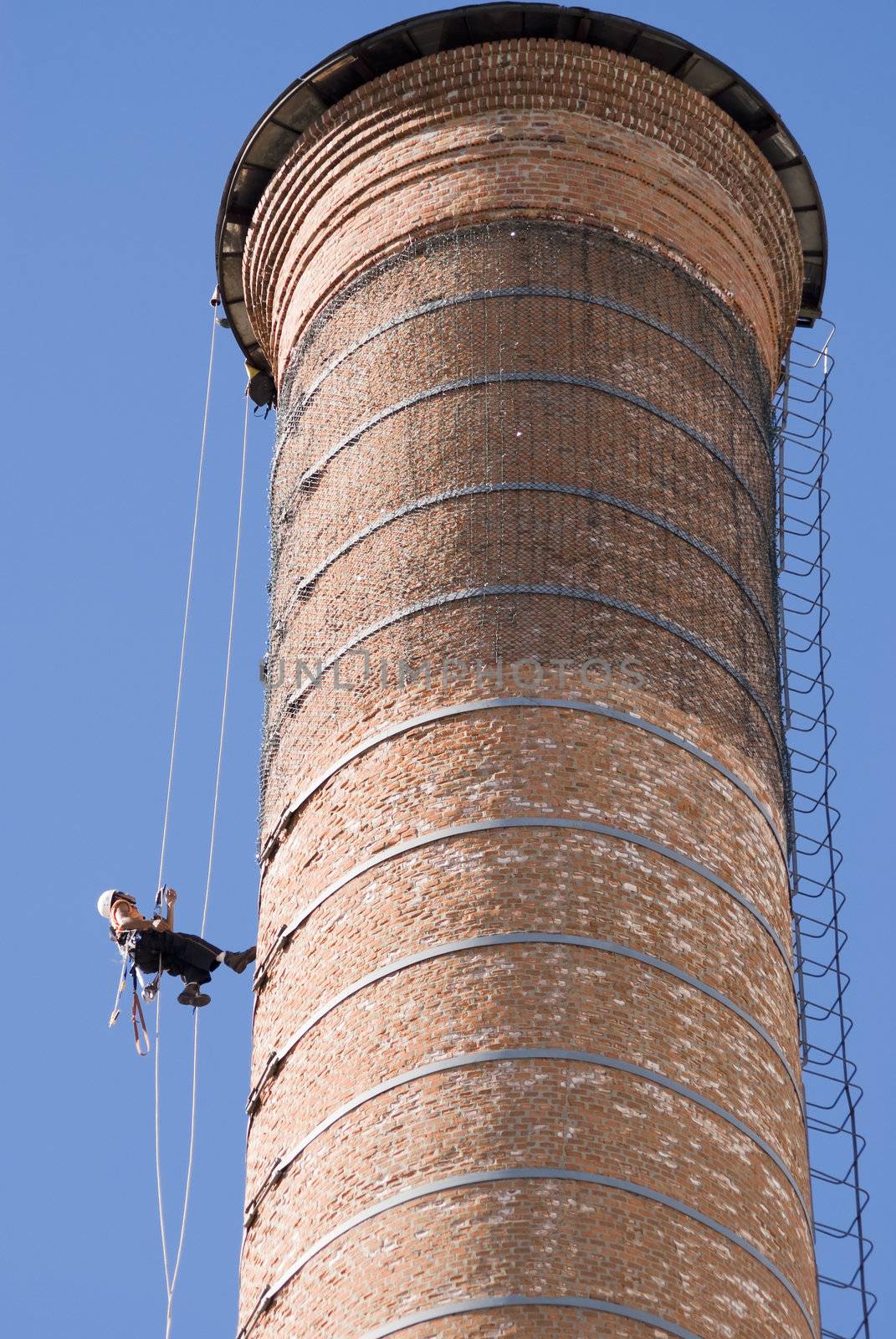 Cropped shot of an unrecognizable workers hanging from a high industrial brick chimney. Blue sky in the background.