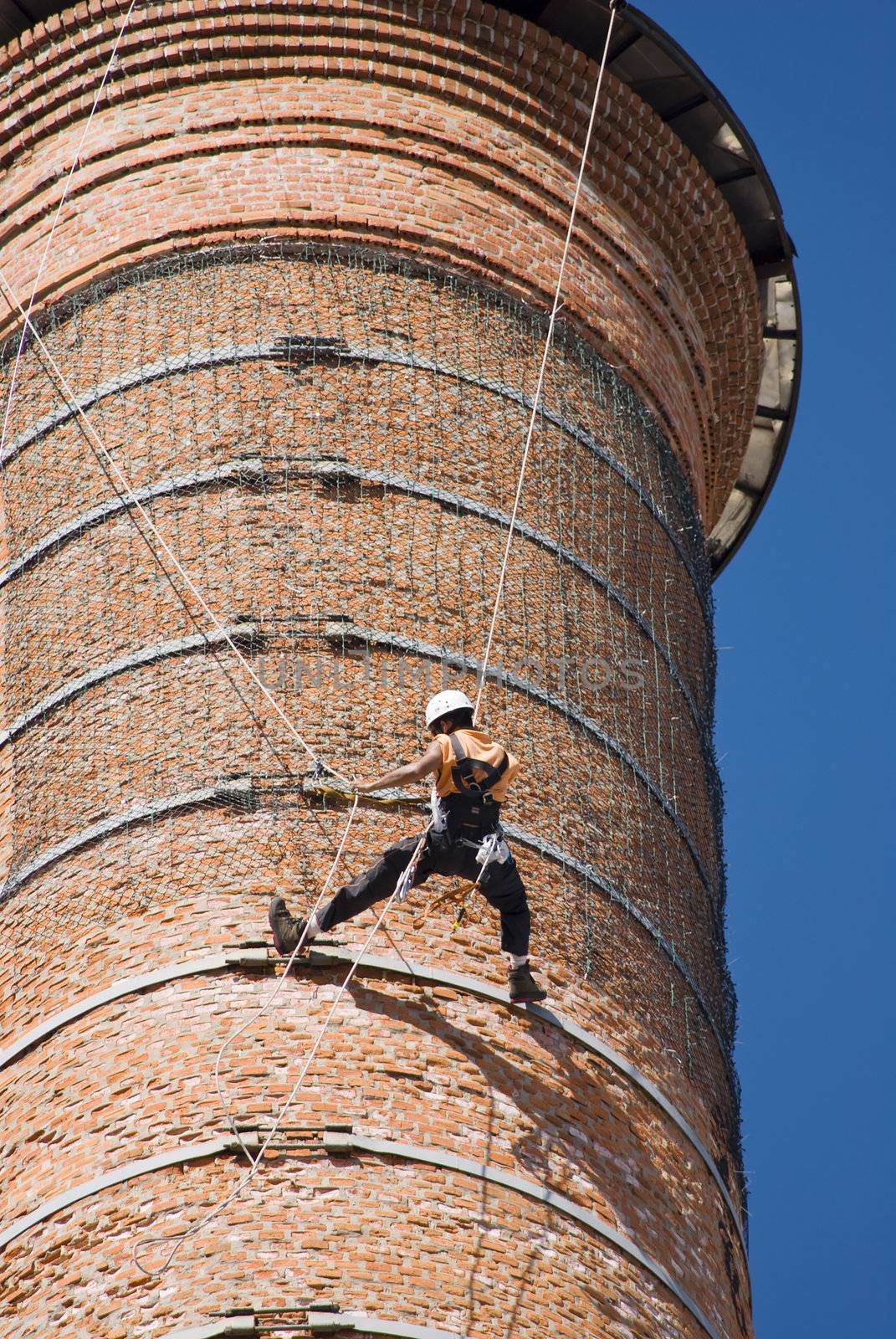 Cropped shot of an unrecognizable workers hanging from a high industrial brick chimney. Blue sky in the background.
