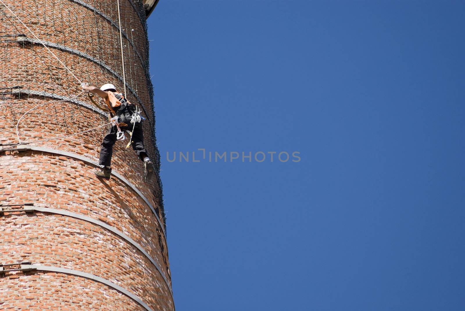 Cropped shot of an unrecognizable workers hanging from a high industrial brick chimney. Blue sky in the background.
