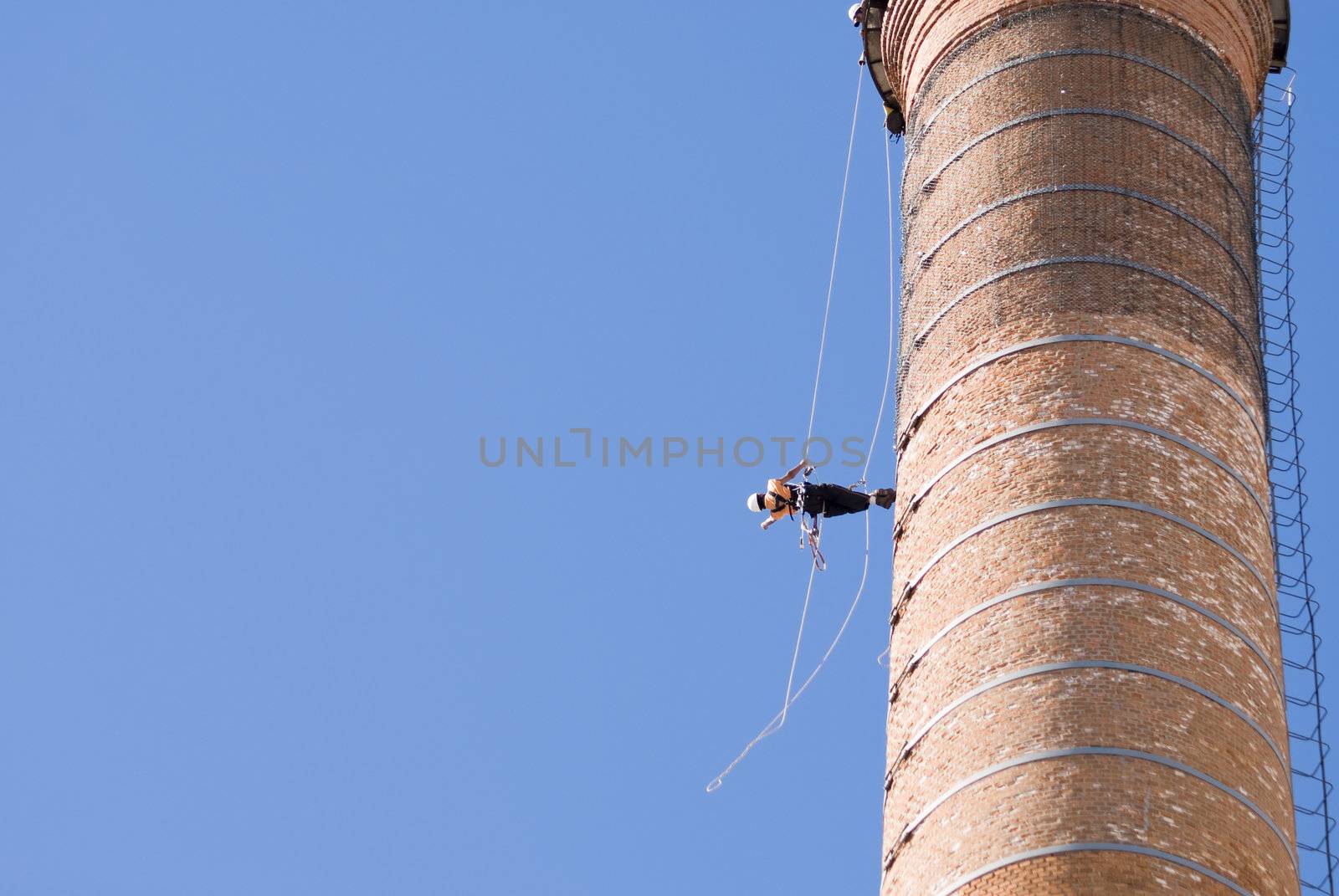 Cropped shot of an unrecognizable workers hanging from a high industrial brick chimney. Blue sky in the background, with huge copy space.