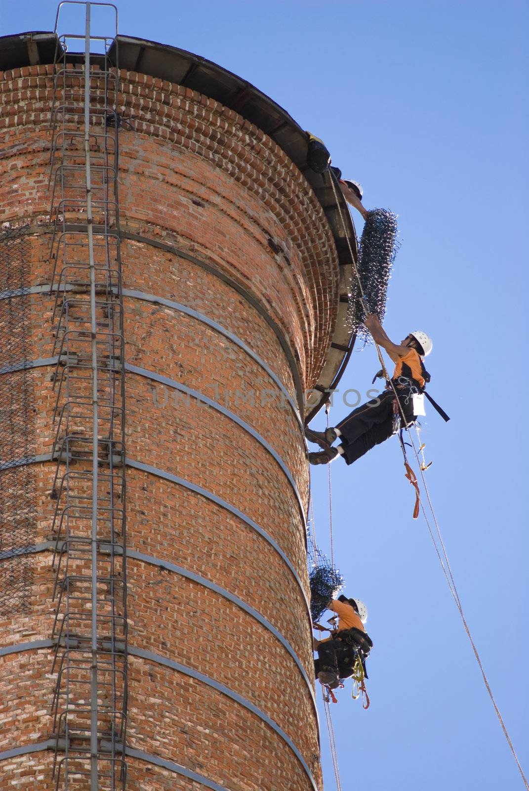 Cropped shot of two unrecognizable workers hanging from a high industrial brick chimney. Blue sky in the background.
