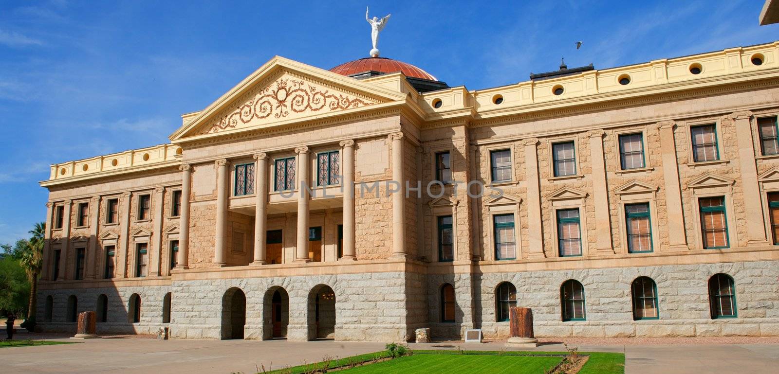 An Arizona State Capital building with circular pillars topped off with a copper dome and white angel with a bright blue sky and green grass.