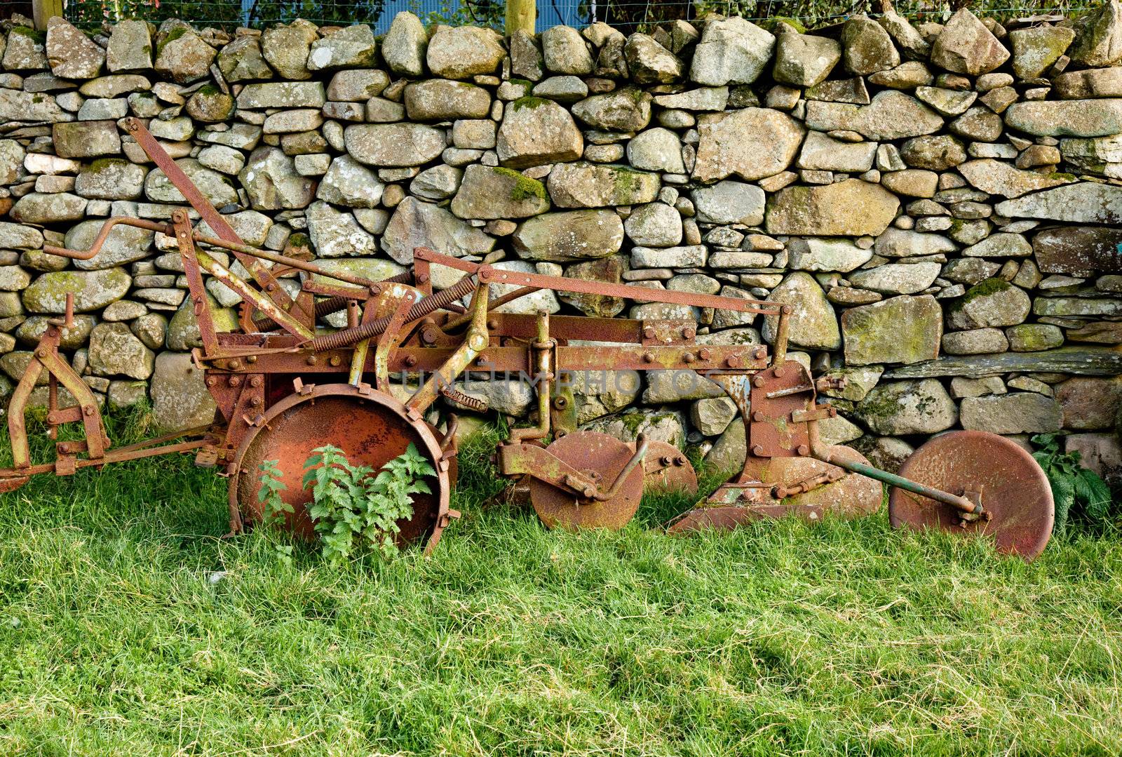 Old rusty plow in shadow of stone wall by steheap