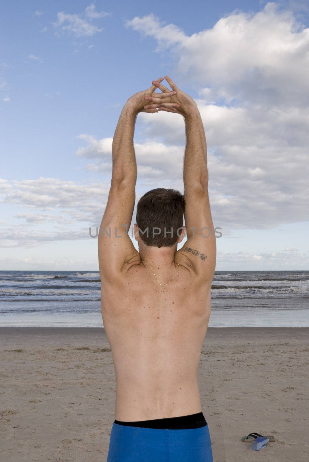 Young man stretching in the beach with cloudy sky. Shot from behind.