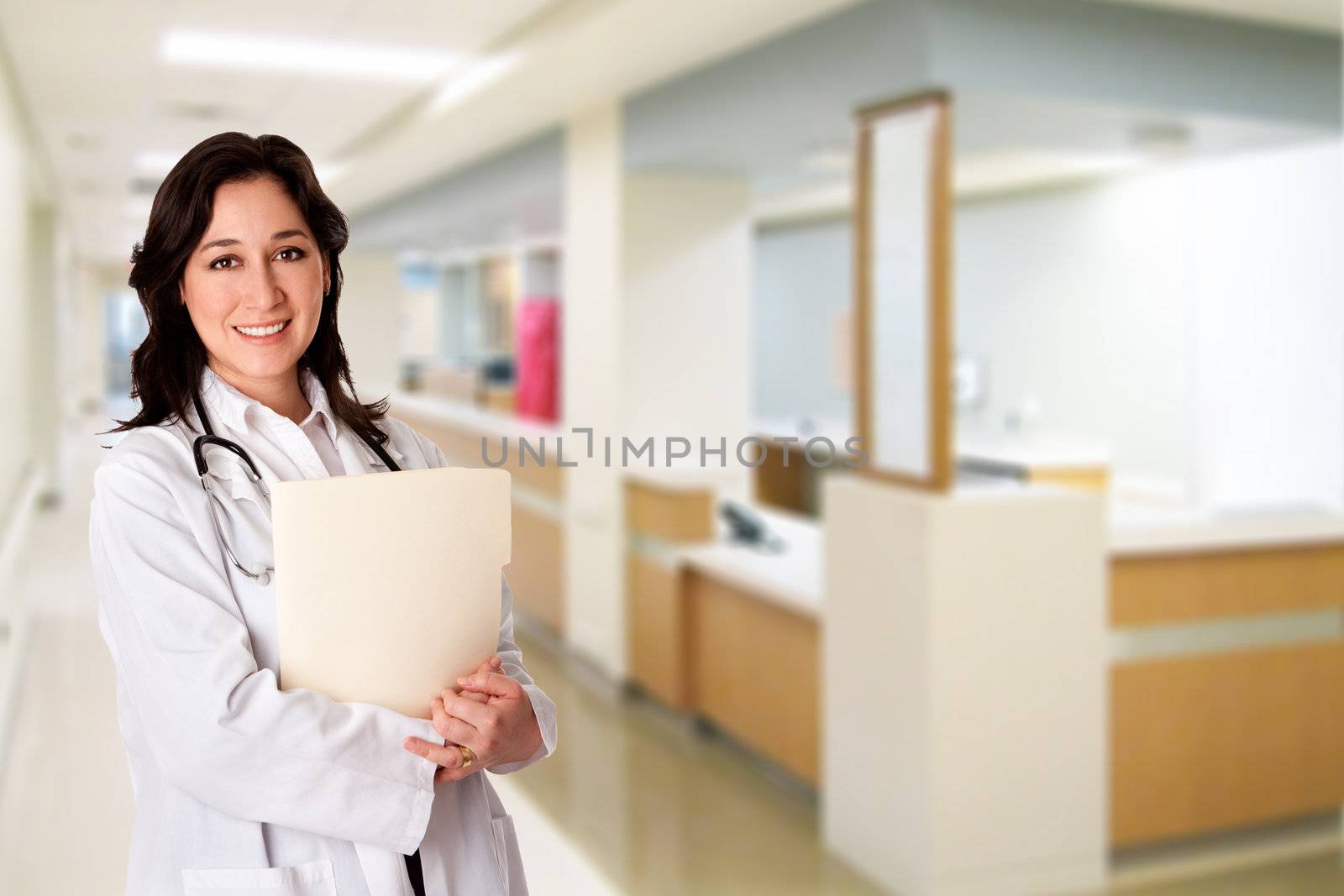 Attractive female doctor with white coat and stethoscope standing holding a patient file chart dossier in corridor hallway at hospital clinic with nursestation desk.