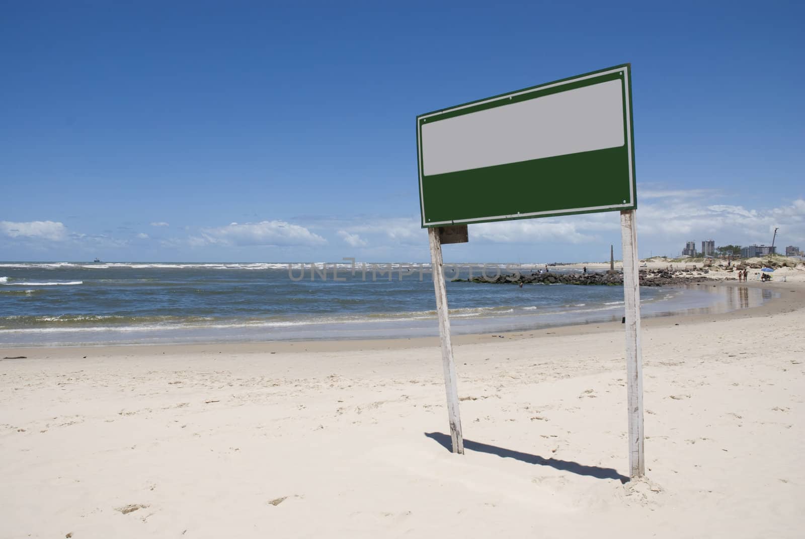 Blank green and white sign board on a beach, with blue sky.