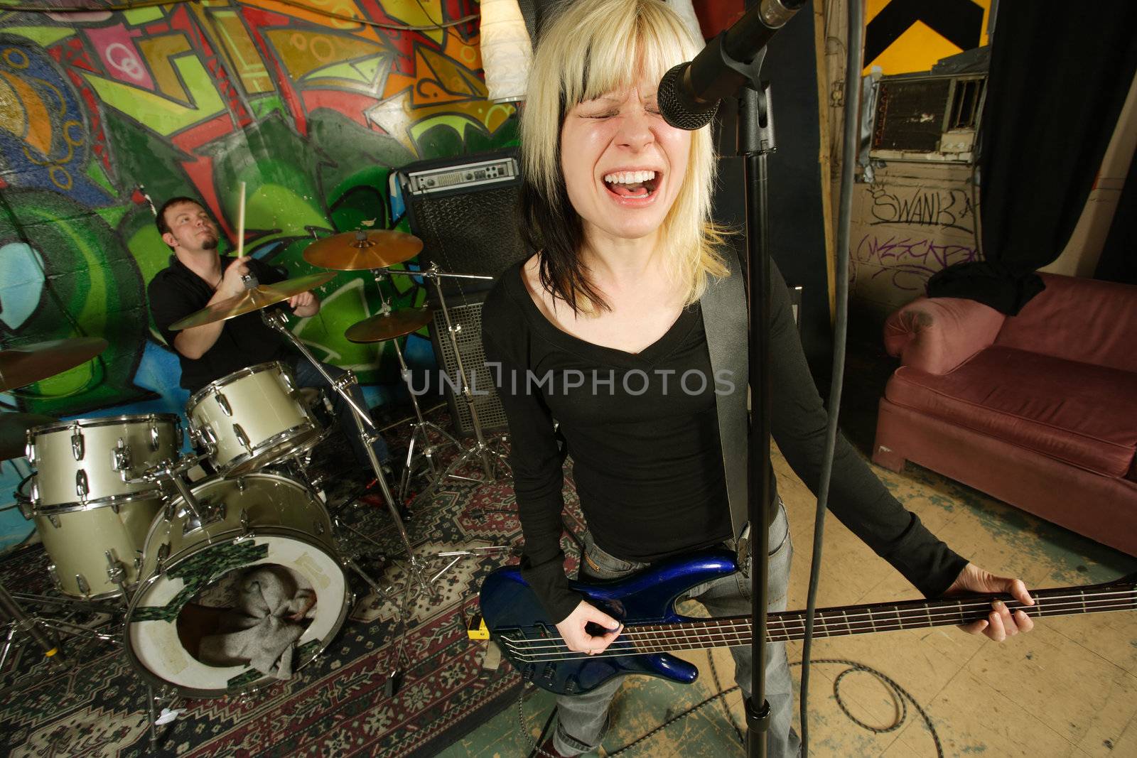 Female bass player screaming into a microphone.  Focus on singers face - drummer blurry in the background.  Shot with slow shutter speed and strobes - motion blur visible in some areas.
