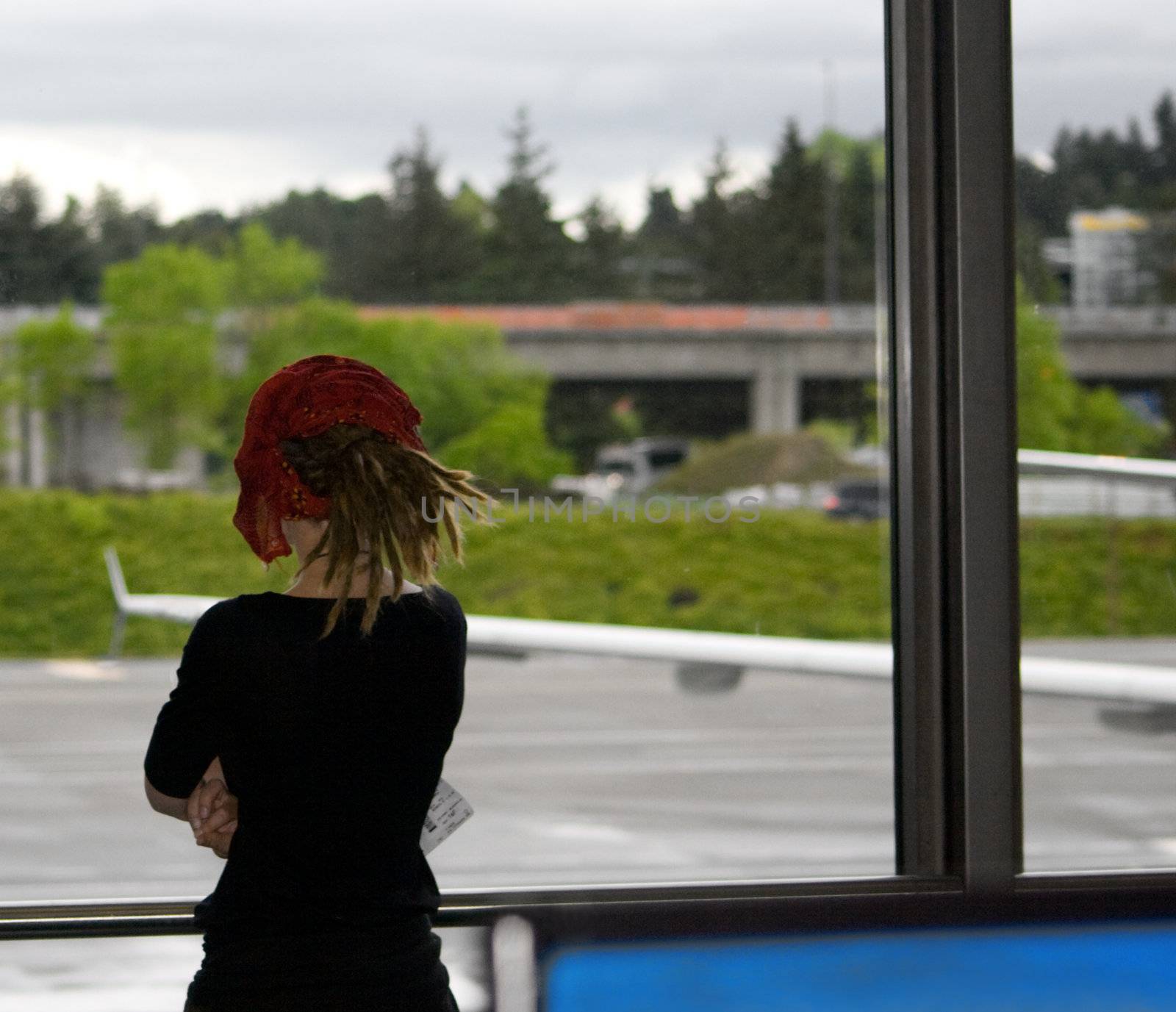 Female Airport Traveler v1 shows a lone young woman looking out an airport terminal window as she waits for her departing flight. She has bags or luggage sitting next to her as she waits. There is an aircraft wing showing of a nearby plane.
