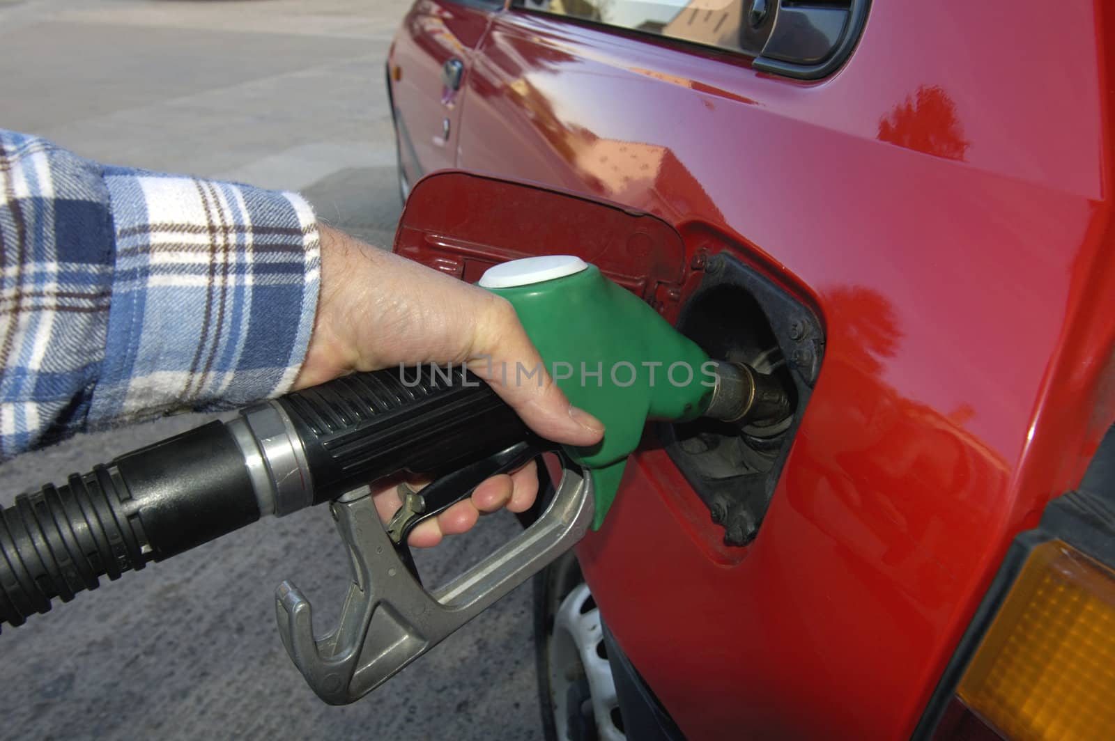 Close up of the hand of a motorist, filling his car with unleaded petrol on a garage forecourt. Beyond is the empty forecourt, but reflected in the car's bodywork is a housing estate and trees.