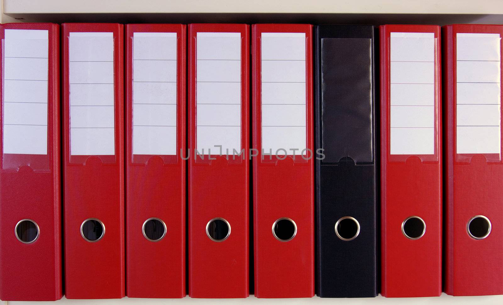 A row of red lever arch files on a shelf, with one black, un-marked one in their midst.