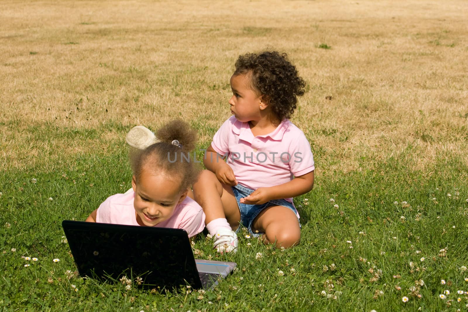 Two Beautiful Ethnic Girls with Laptop Computer in the park on a field of clovers and brown and green grass.