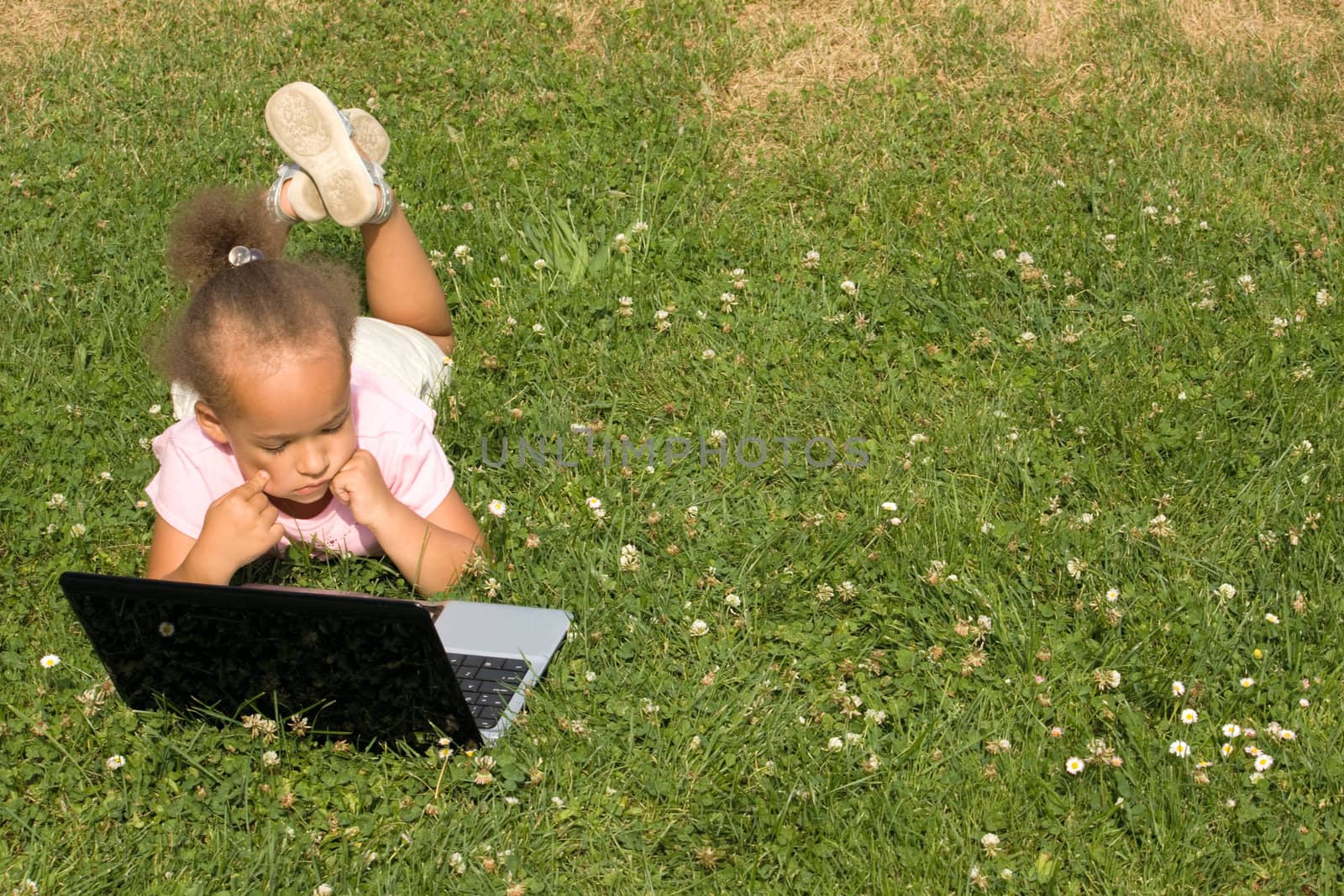 Beautiful young ethnic race girl using laptop on a field of green grass and daisy wheel and clovers. She has the expression of contemplating as she surfs the internet.