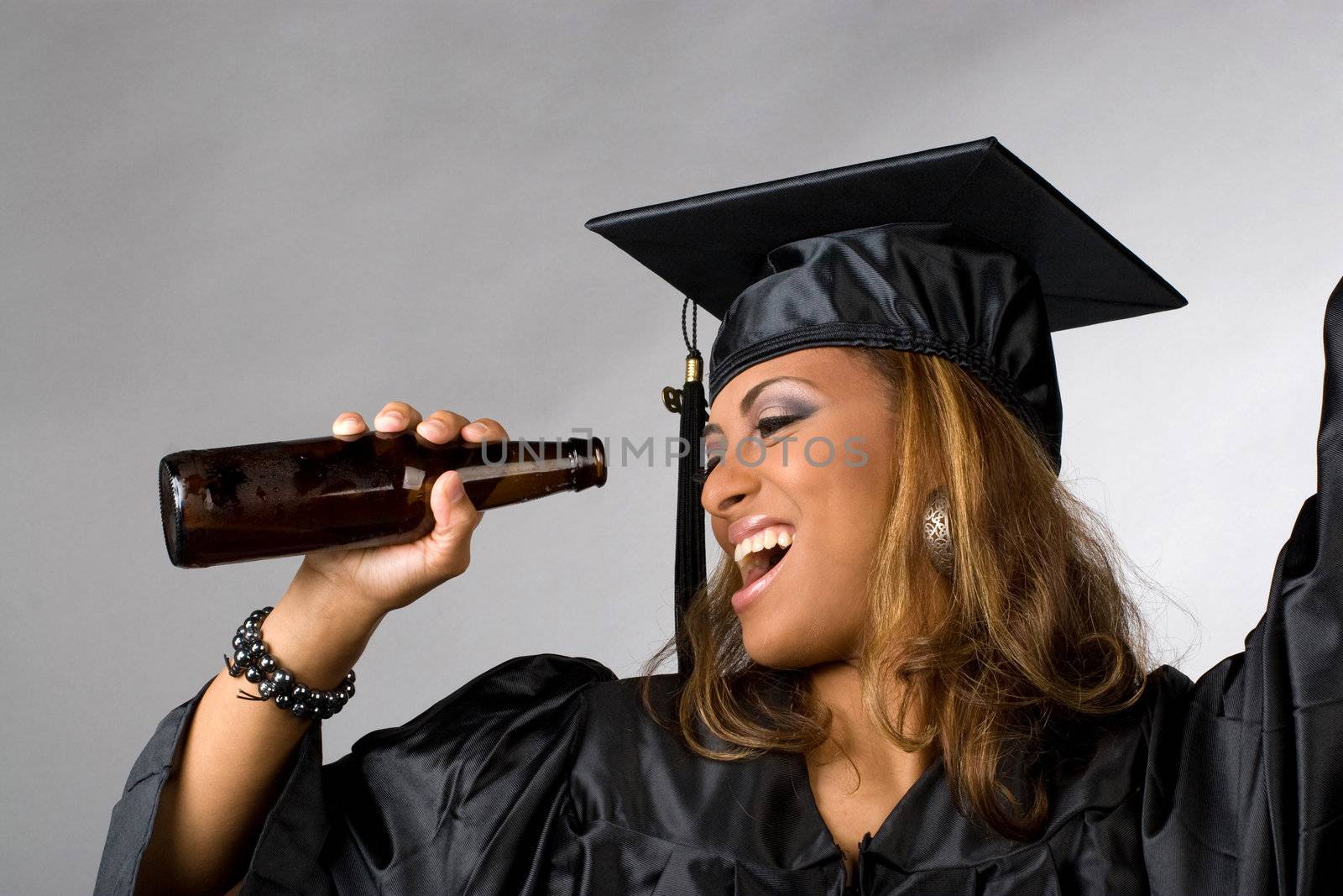 A recent graduate posing in her cap and gown holding beer bottle isolated over a silver background.