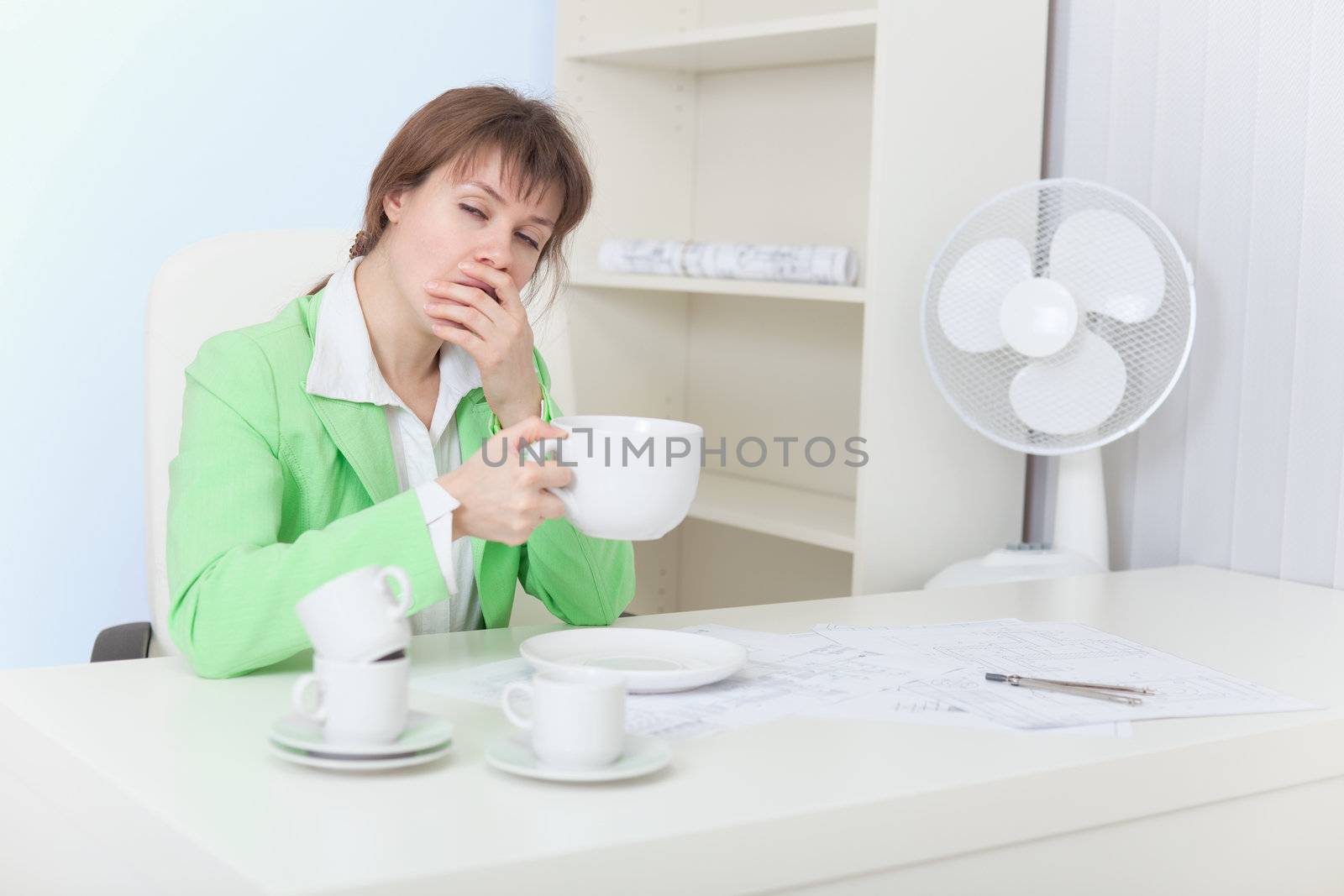 The tired woman sits at a table with the big cup of coffee and yawns