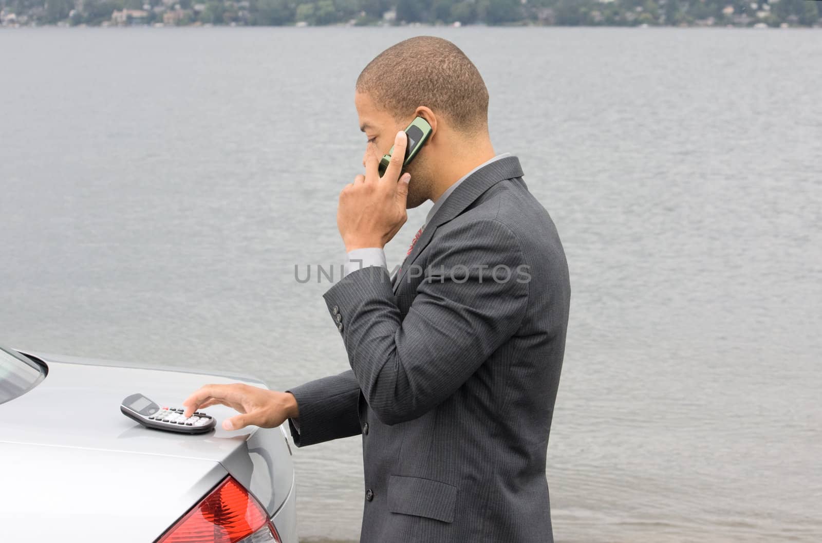 Ethnic Business Man using calculator while talking on cell phone. He is parked at the shore of the lake.