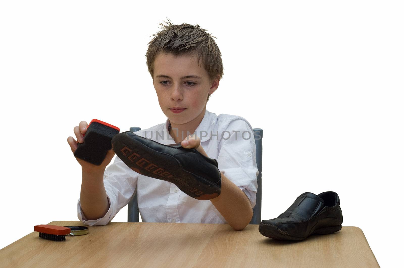 a young teenage boy sat at the table polishing his school shoes using a brush and buffing sponge.